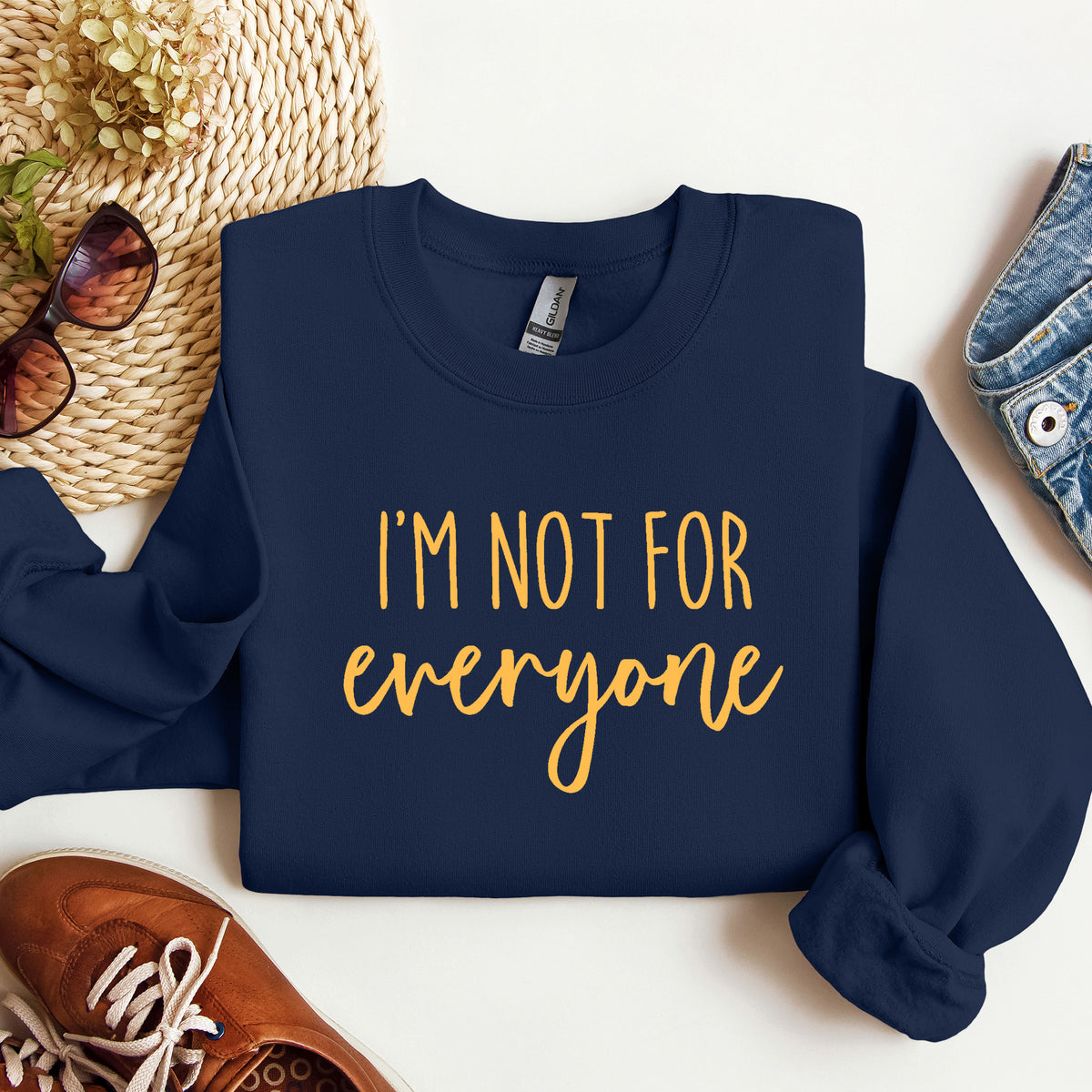 A navy blue crewneck sweatshirt with the text 'I'M NOT FOR everyone' printed in gold on the front, styled in a flat lay with jeans, sunglasses, a woven hat, and brown sneakers on a white background.