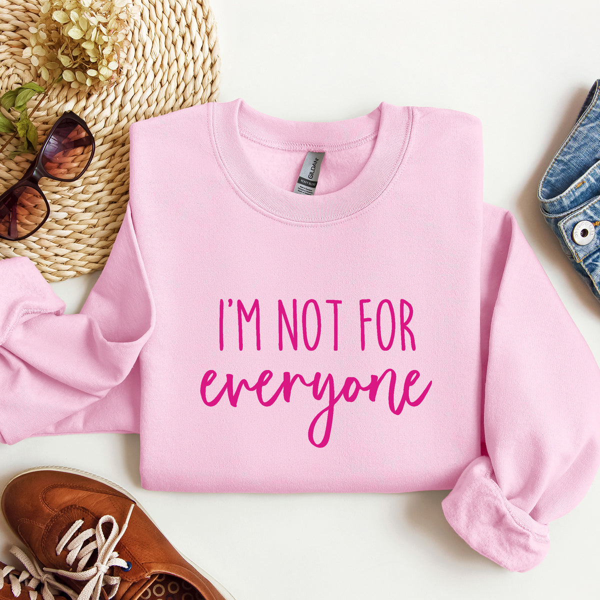 A folded light pink crewneck sweatshirt with the text 'I'M NOT FOR everyone' in pink lettering, styled in a flat lay with sunglasses, a woven hat, a brown shoe, and a denim item on a white background.