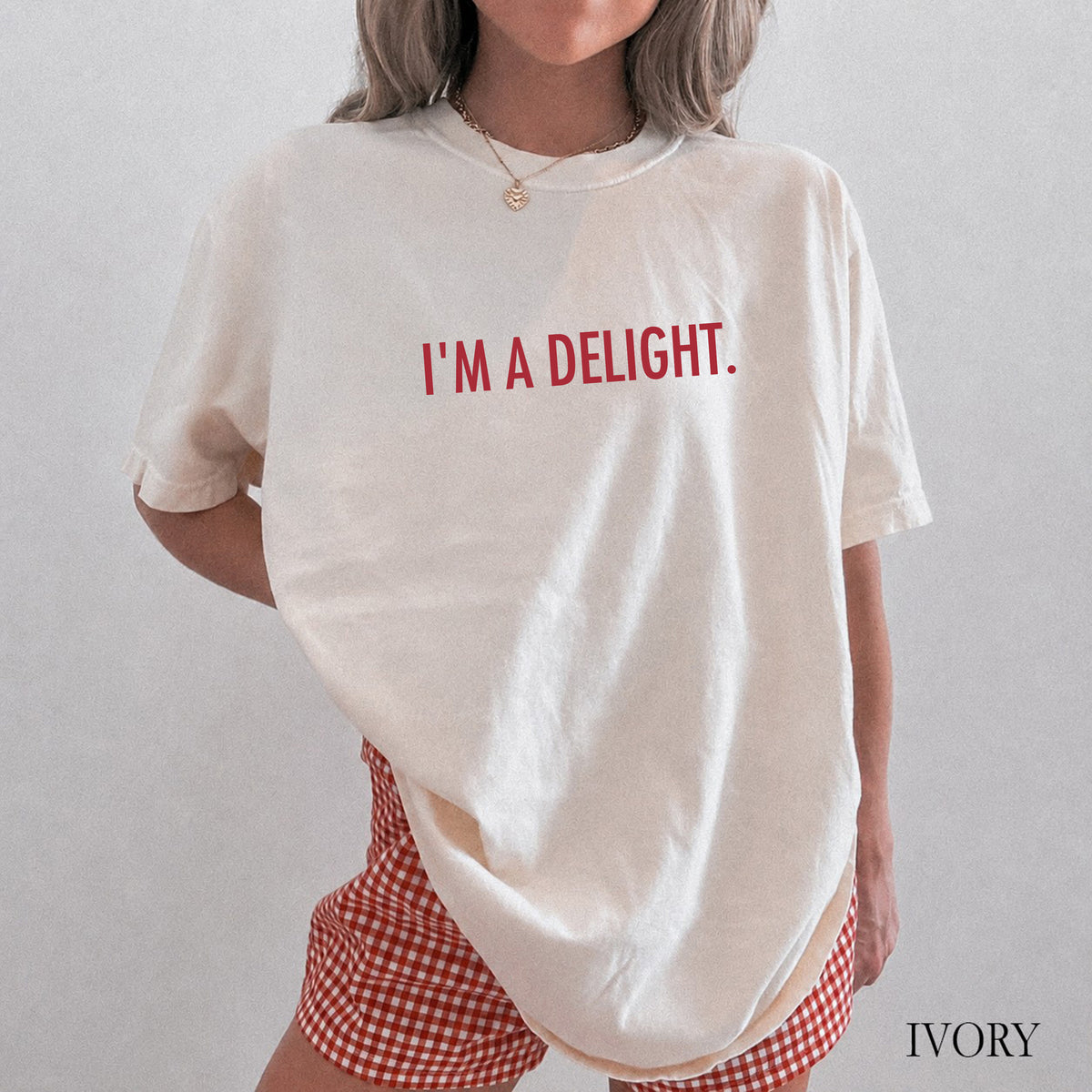 A person wearing an oversized ivory T-shirt with the phrase 'I'M A DELIGHT.' printed in red across the chest, paired with red-and-white gingham shorts.