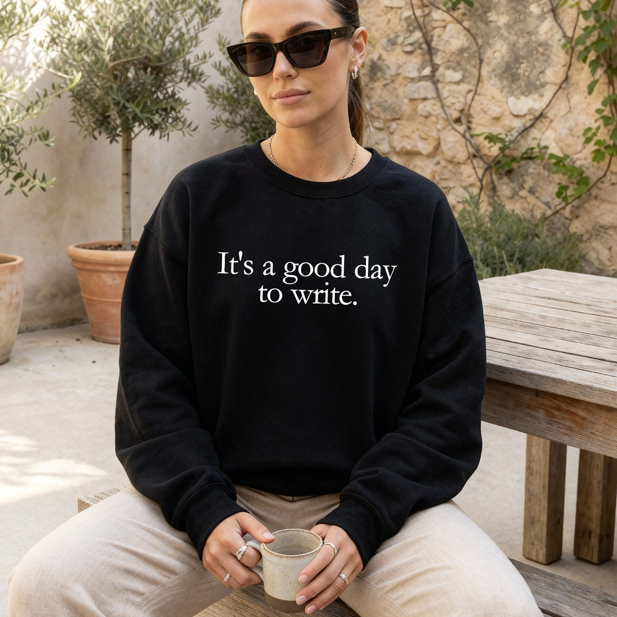 A woman wearing a black crewneck sweatshirt with the white printed text 'It's a good day to write.' sits outdoors on a wooden bench, holding a ceramic mug.