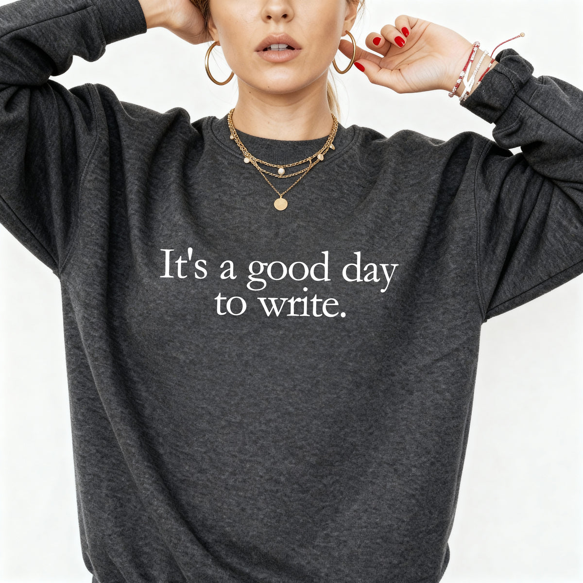 A woman wearing a dark heather gray crewneck sweatshirt with the white printed text 'It's a good day to write.' on the chest, styled with gold jewelry against a plain white background.