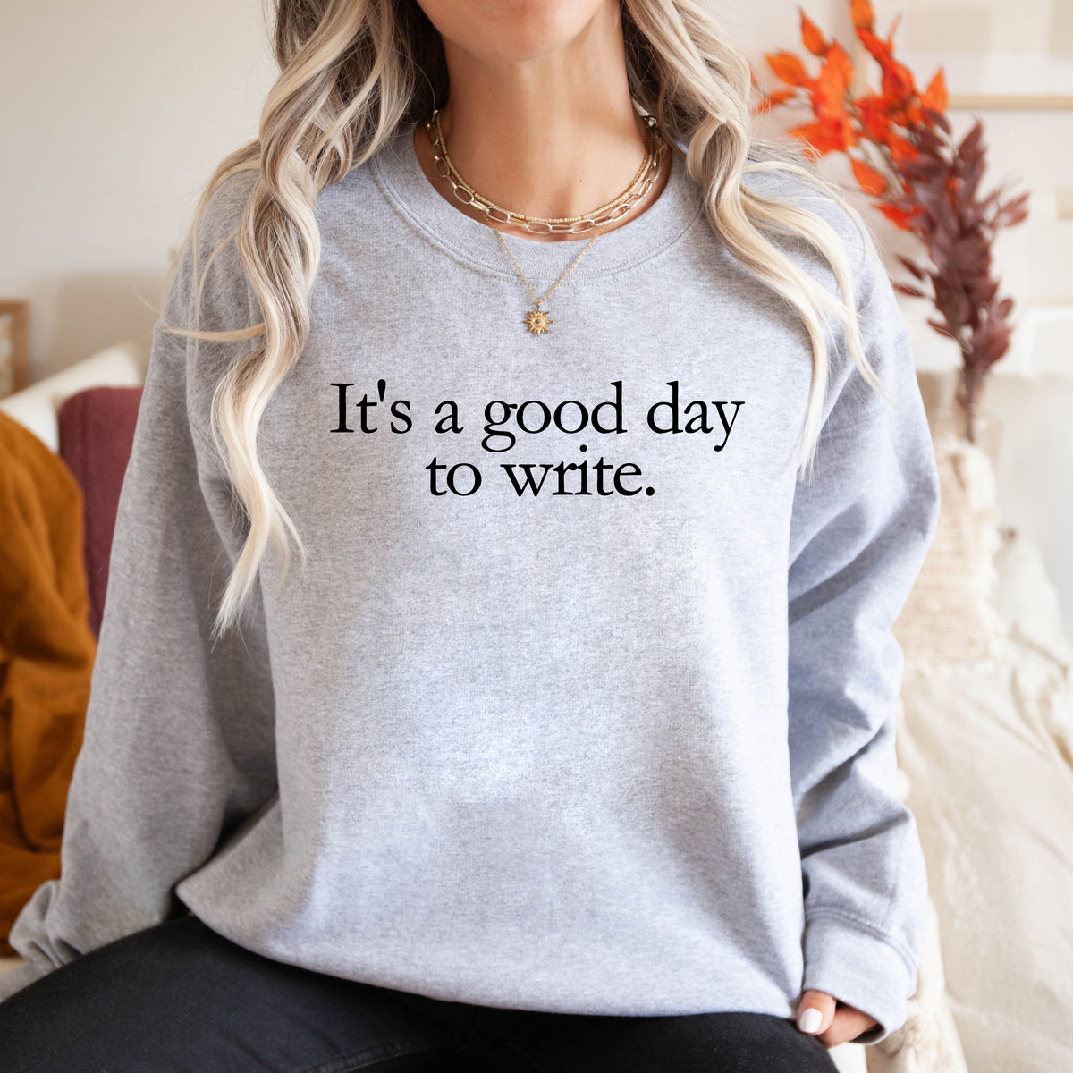 A woman wearing a light heather gray crewneck sweatshirt with the black printed text 'It's a good day to write.' on the chest, styled with layered gold necklaces in a cozy indoor setting.