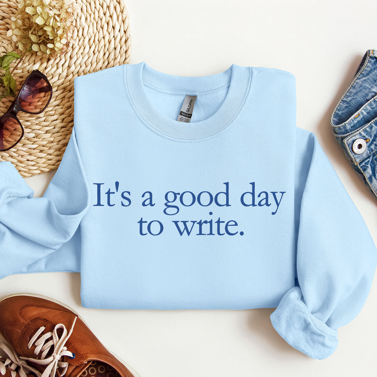 A light blue crewneck sweatshirt with the phrase 'It's a good day to write.' printed in navy blue serif text across the chest.