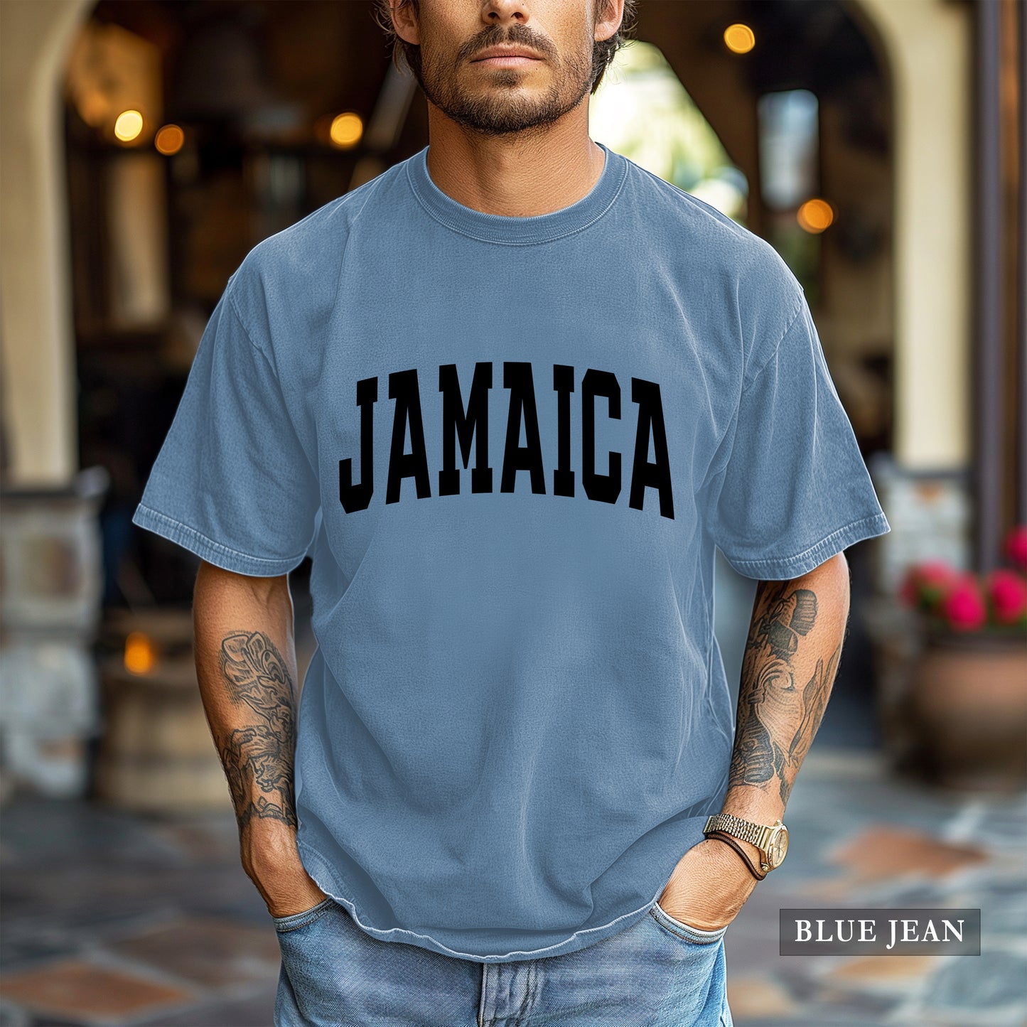 A man wearing a light blue crewneck t-shirt with 'JAMAICA' printed in bold black letters across the chest, set against a blurred outdoor patio background.