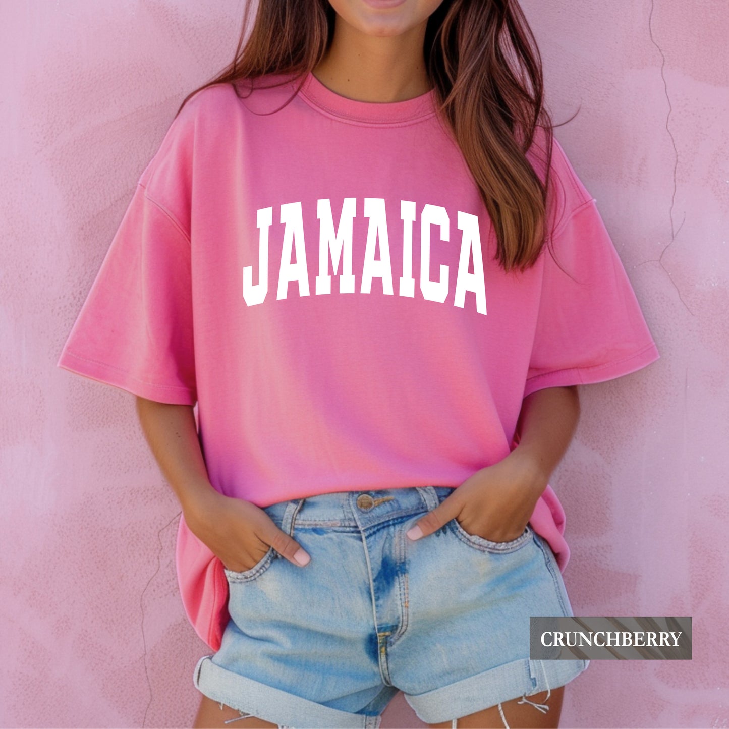 A woman wearing a pink oversized t-shirt with 'JAMAICA' printed in white arched letters across the chest, paired with light blue denim shorts, standing against a textured pink wall. A 'CRUNCHBERRY' brand label is visible in the bottom right corner.