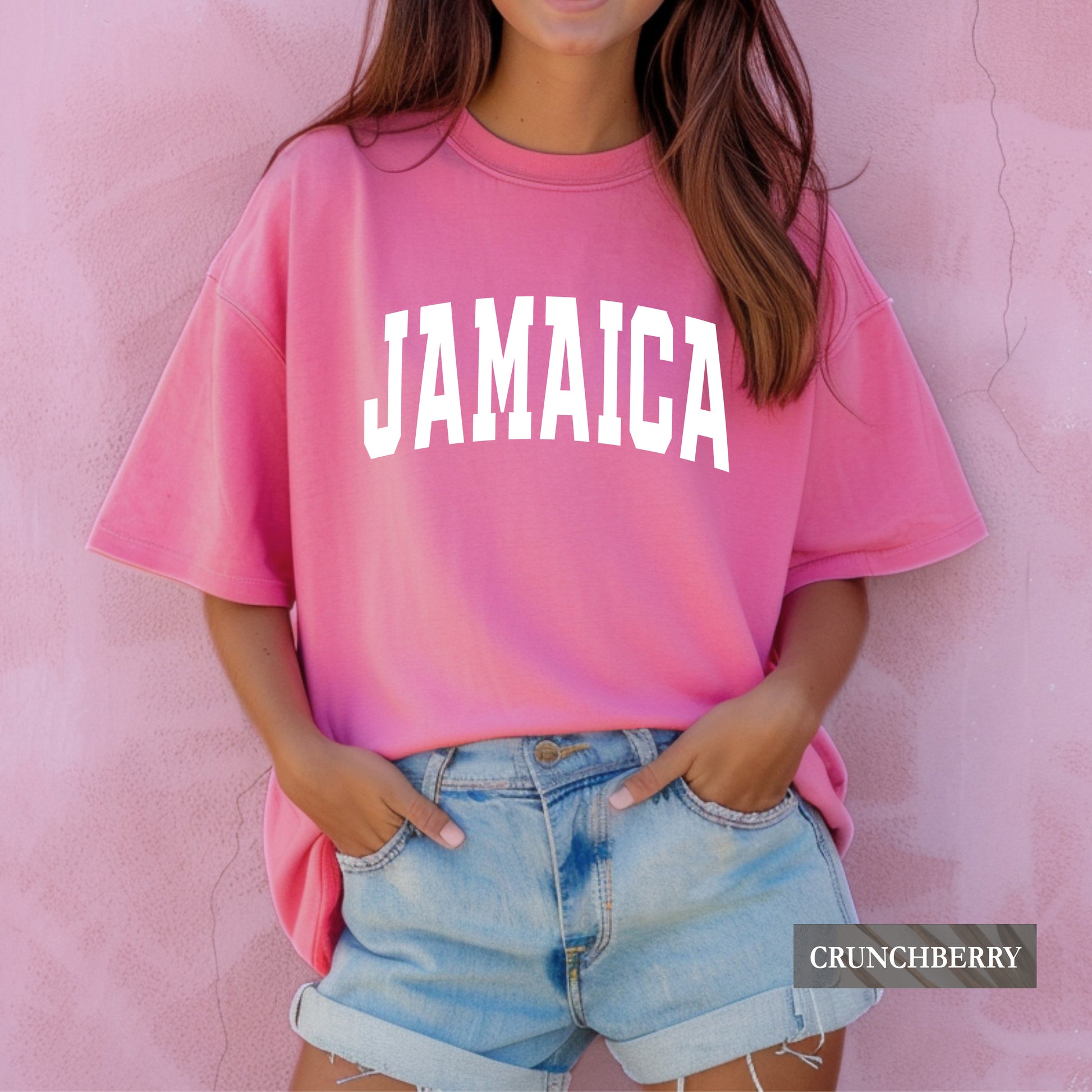 A woman wearing a pink oversized t-shirt with 'JAMAICA' printed in white arched letters across the chest, paired with light blue denim shorts, standing against a textured pink wall. A 'CRUNCHBERRY' brand label is visible in the bottom right corner.
