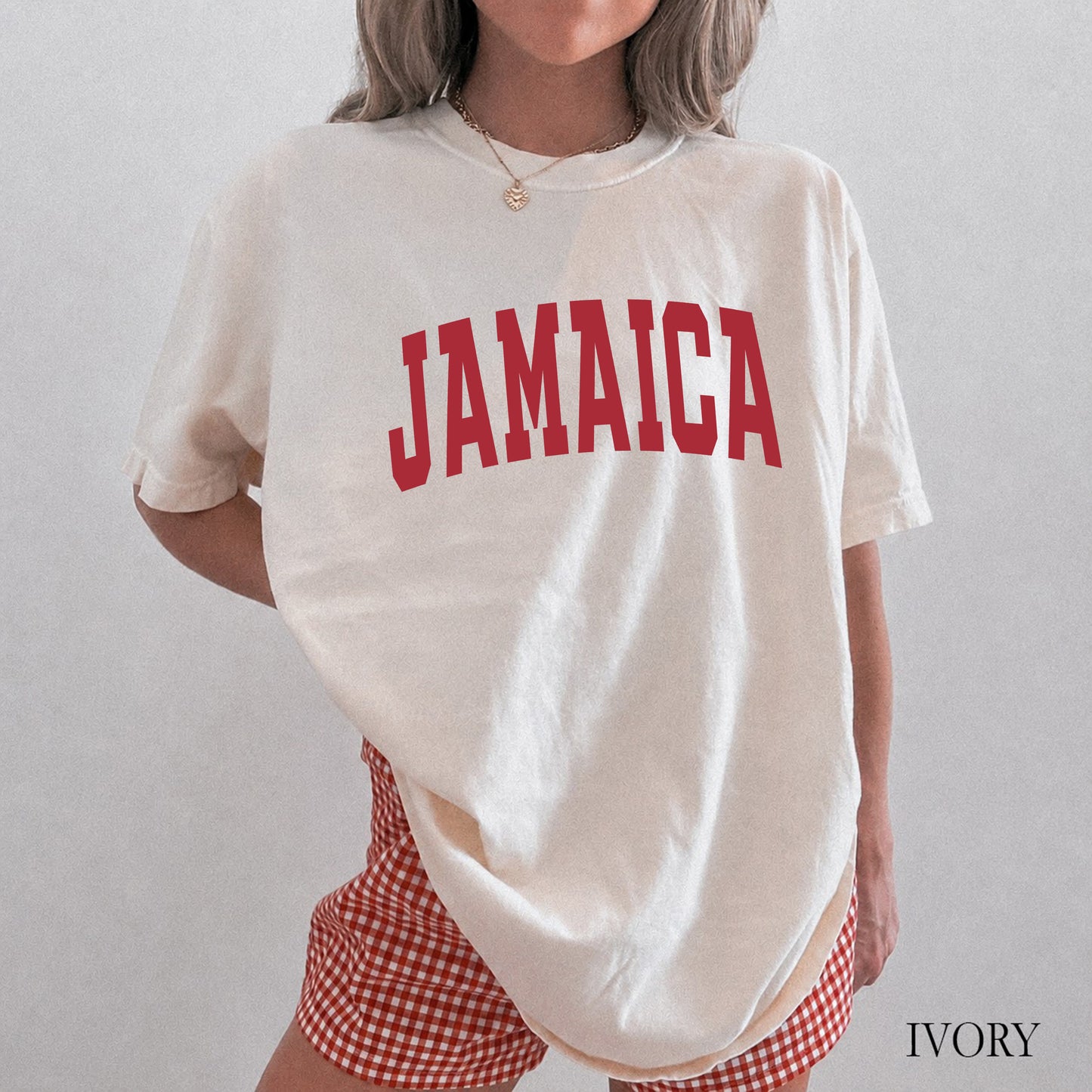 A woman wearing an oversized cream-colored t-shirt with 'JAMAICA' printed in red arched letters across the chest, paired with red and white gingham shorts. The image has a text overlay 'IVORY' in the bottom right corner.