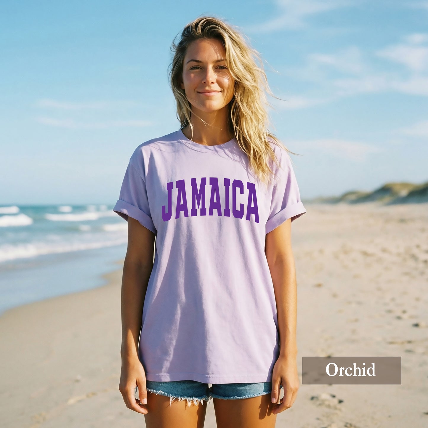 A woman standing on a sandy beach wearing an oversized light orchid purple t-shirt with 'JAMAICA' printed in dark purple across the chest, paired with denim shorts. A text overlay in the bottom right corner reads 'Orchid'.