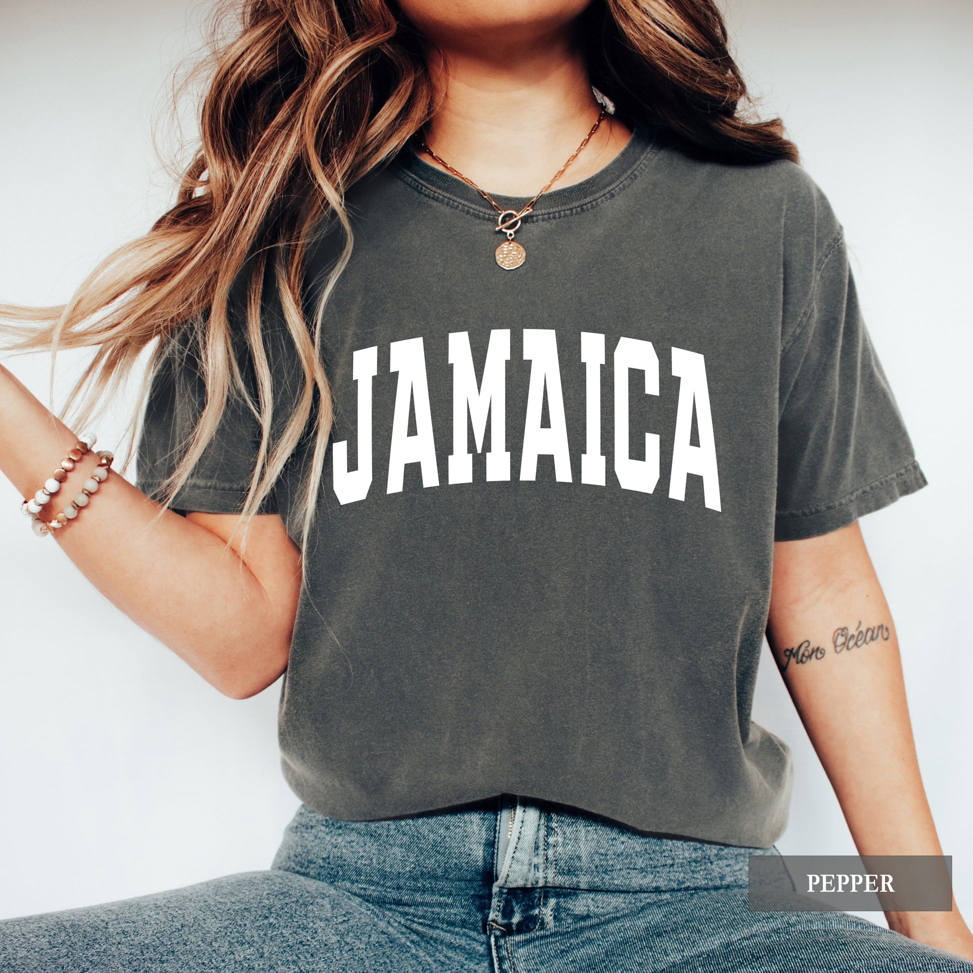 A woman wearing a washed charcoal gray t-shirt with 'JAMAICA' printed in bold white letters across the chest, paired with blue jeans. The image has a 'PEPPER' color label overlay in the bottom right corner.