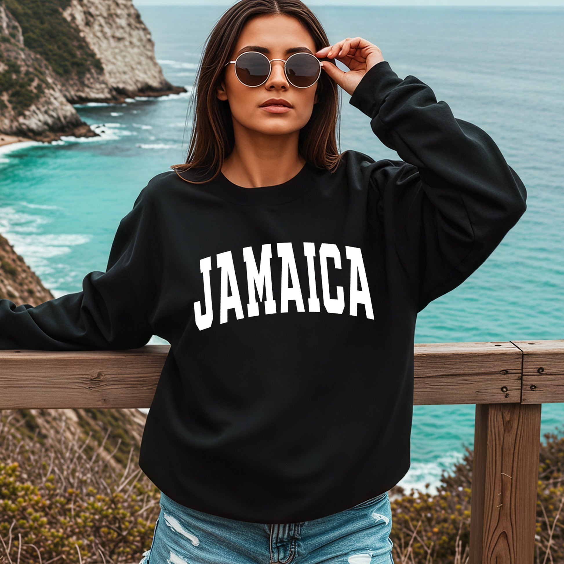 A woman wearing a black crewneck sweatshirt with 'JAMAICA' printed in bold white varsity letters across the chest, standing on a wooden railing with a scenic ocean and rocky coastline in the background.