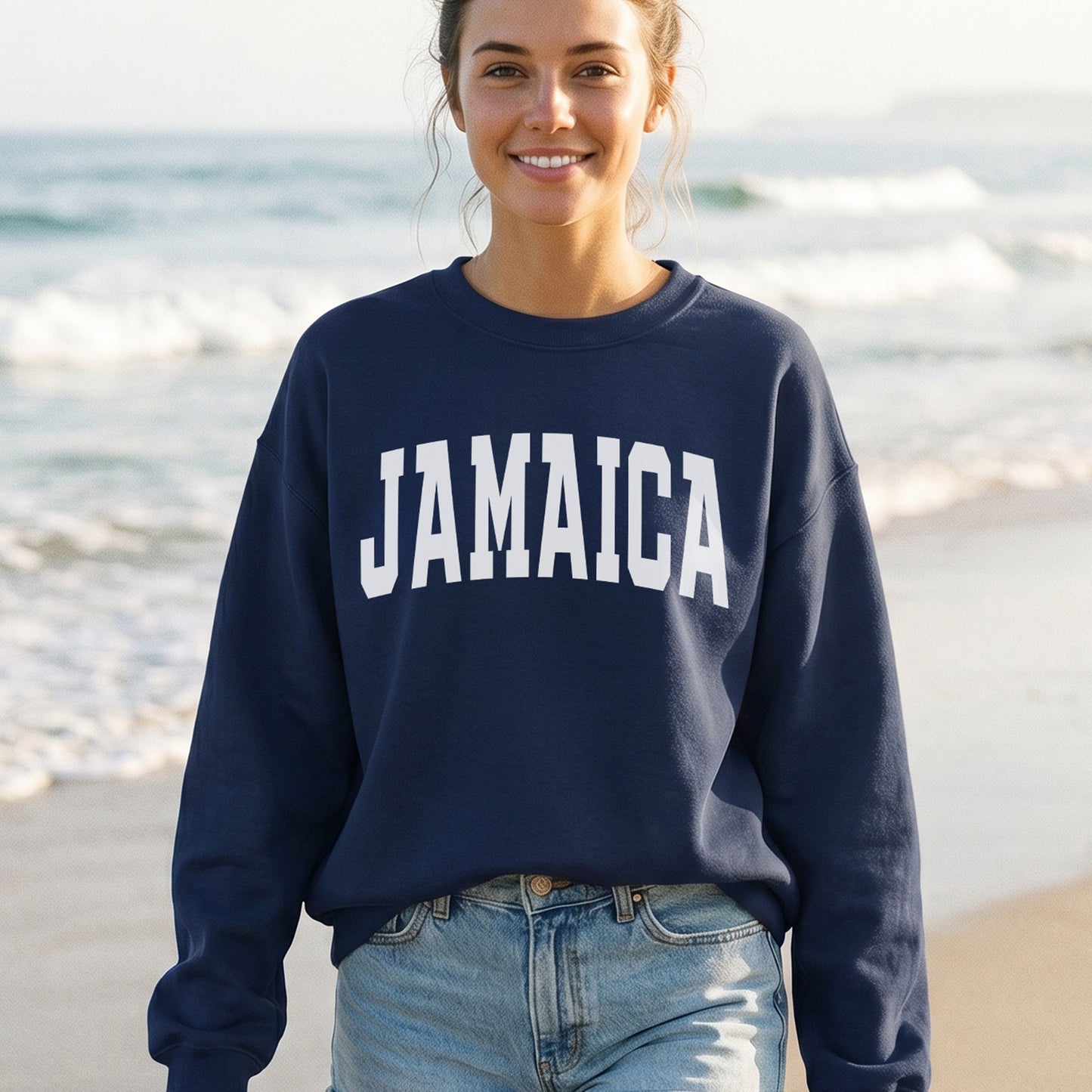 A woman standing on a beach wearing a navy blue sweatshirt with 'JAMAICA' printed in bold white letters across the chest, paired with light blue jeans.