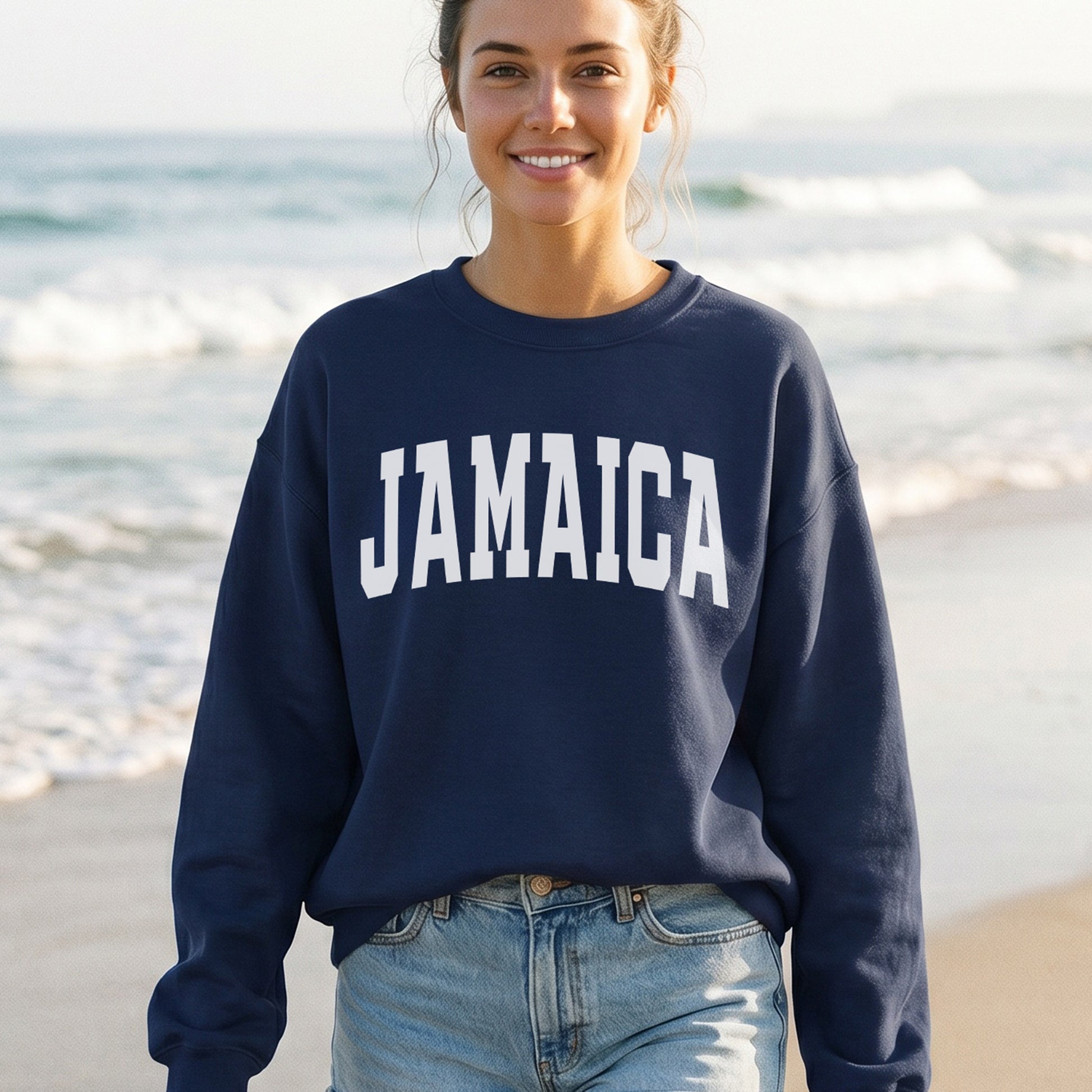 A woman standing on a beach wearing a navy blue sweatshirt with 'JAMAICA' printed in bold white letters across the chest, paired with light blue jeans.