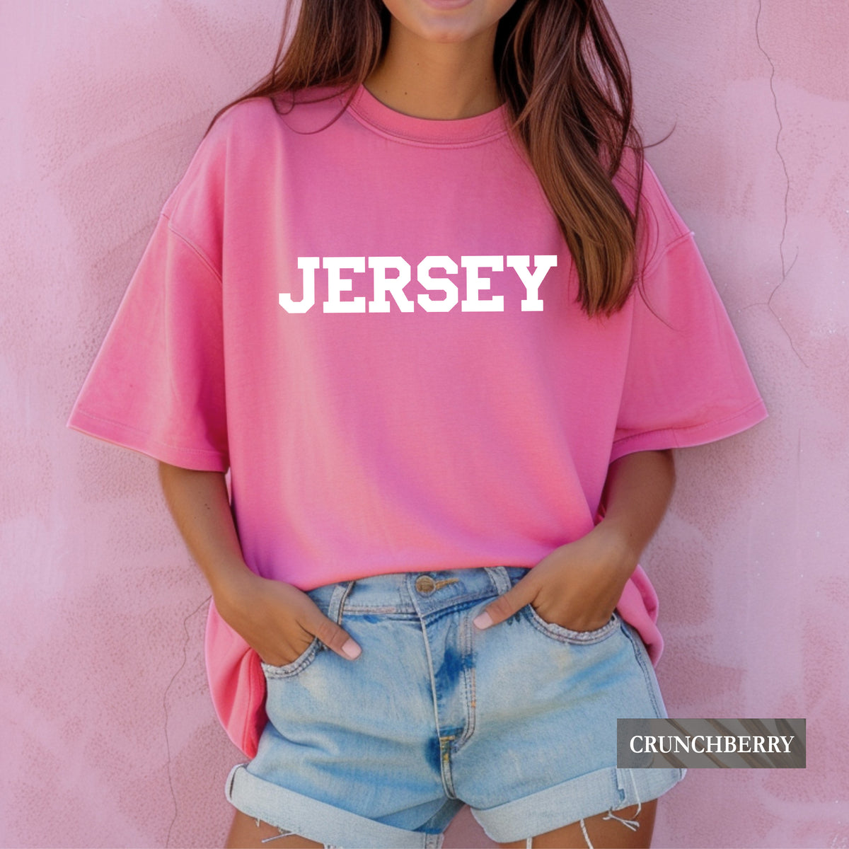 A woman wearing a pink oversized crewneck t-shirt with the word 'JERSEY' printed in bold white letters across the chest, paired with light blue denim shorts, standing against a textured pink wall. A 'CRUNCHBERRY' brand label is visible in the bottom right corner.