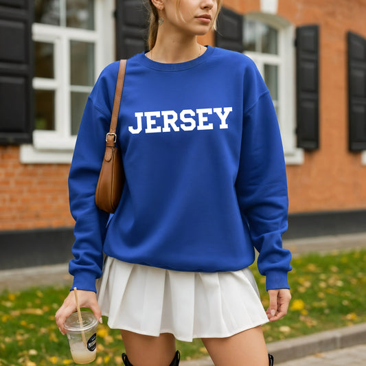A woman wearing a royal blue crewneck sweatshirt with 'JERSEY' printed in white block letters across the chest, paired with a white pleated mini skirt and a brown shoulder bag, standing outdoors in front of a brick building.