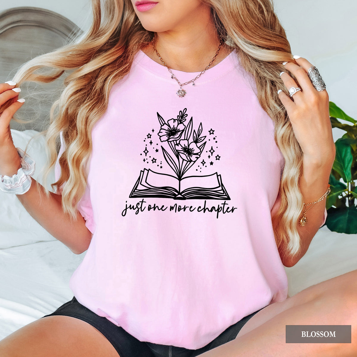A woman wearing a light pink short-sleeve t-shirt with a black graphic of an open book and flowers, with the text 'just one more chapter' printed below. The image is labeled 'BLOSSOM' in the bottom right corner.