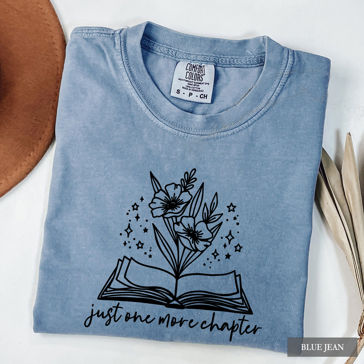 A folded light blue graphic t-shirt in a 'Blue Jean' wash by Comfort Colors, featuring a black line art design of flowers blooming from an open book with the text 'just one more chapter'. The shirt is styled on a white background with a brown leather object on the left and dried leaves on the right.