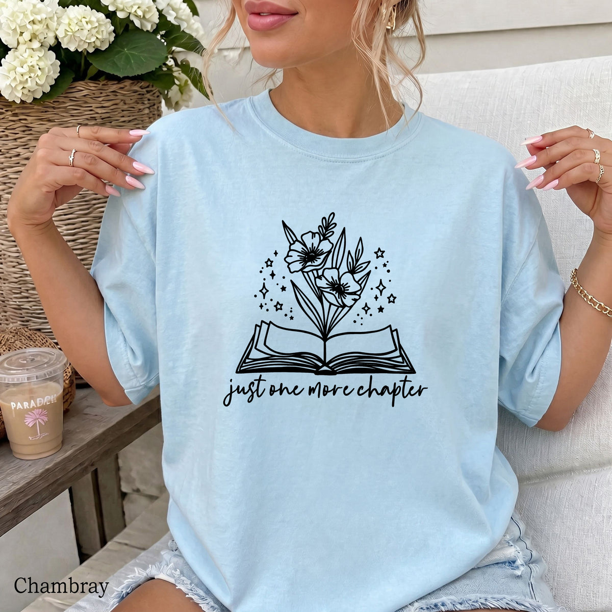 A woman wearing a light chambray blue graphic t-shirt with a black illustration of flowers growing from an open book and the text 'just one more chapter'.