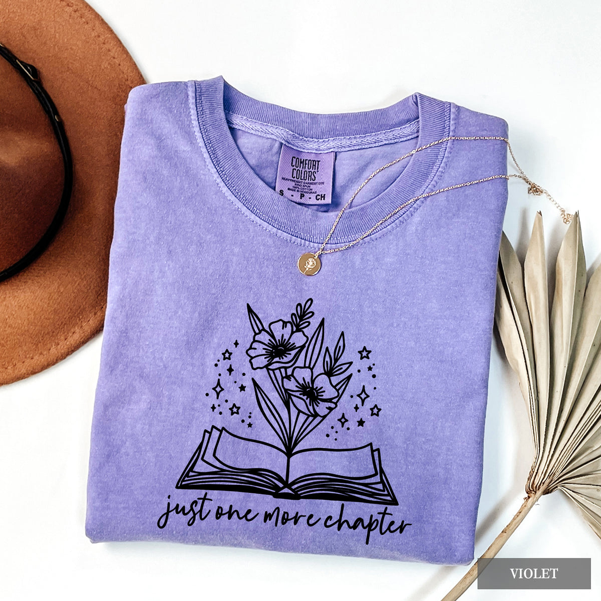 A folded lavender-purple t-shirt with a black graphic of an open book and flowers, styled on a white background with a brown hat, a dried palm leaf, and a gold necklace.