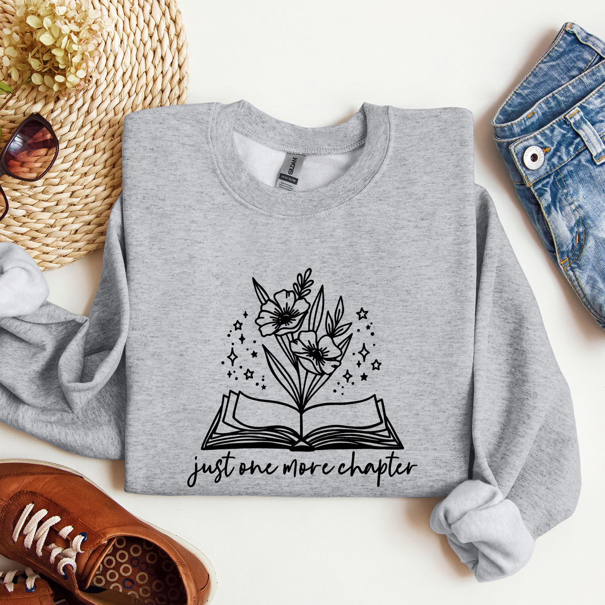 A heather gray crewneck sweatshirt with a black graphic of an open book, flowers, and the text 'just one more chapter', styled in a flat lay with jeans, sunglasses, a straw hat, and brown shoes on a white background.