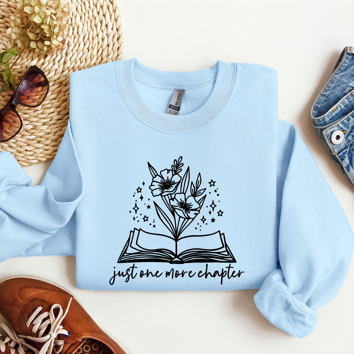 A light blue long-sleeve sweatshirt with a black graphic of an open book, flowers, and stars, and the text 'just one more chapter' printed on the front.