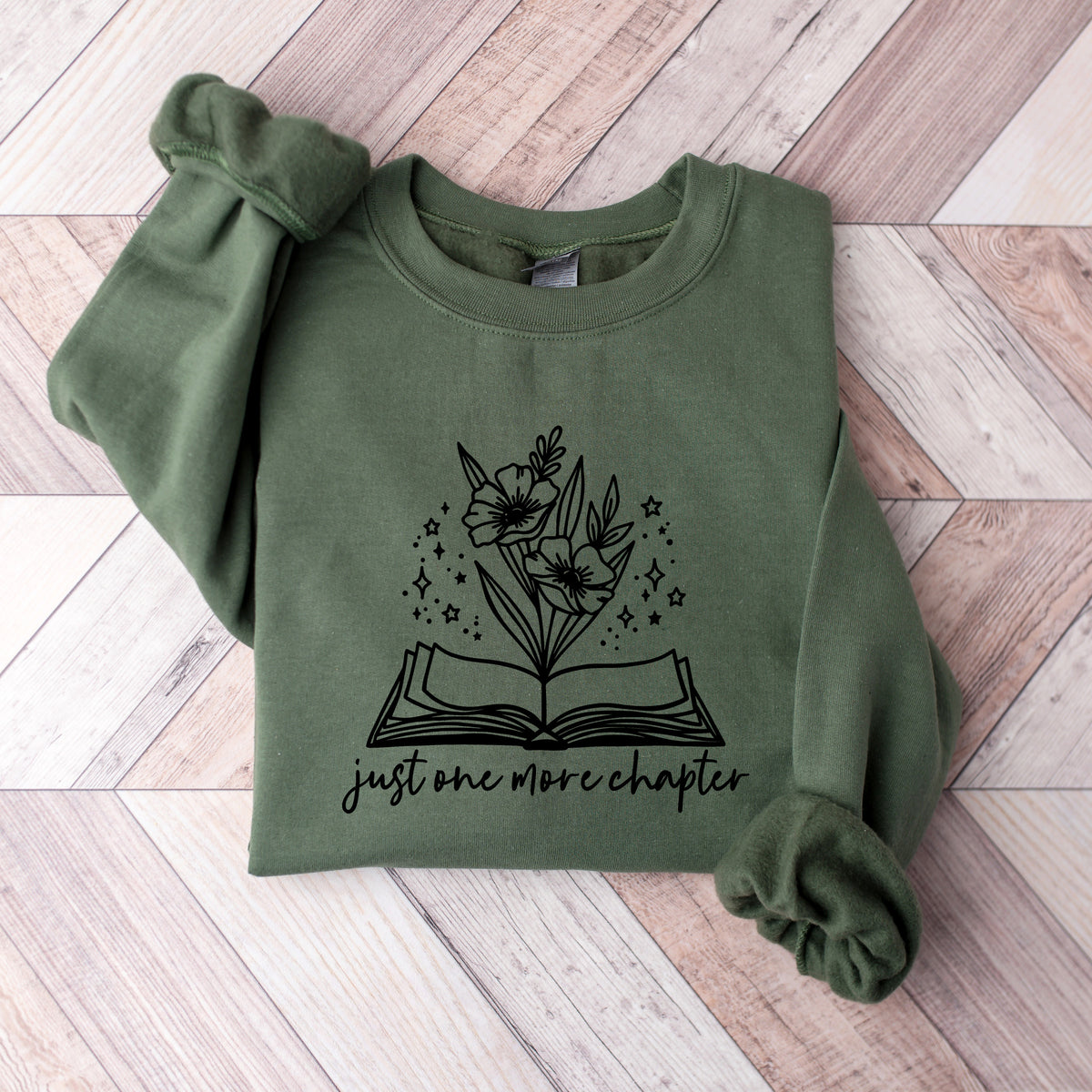 An olive green long-sleeve sweatshirt with a black graphic of an open book, flowers, and stars, and the text 'just one more chapter' printed on the front. The sweatshirt is laid flat on a light-colored wooden plank background.