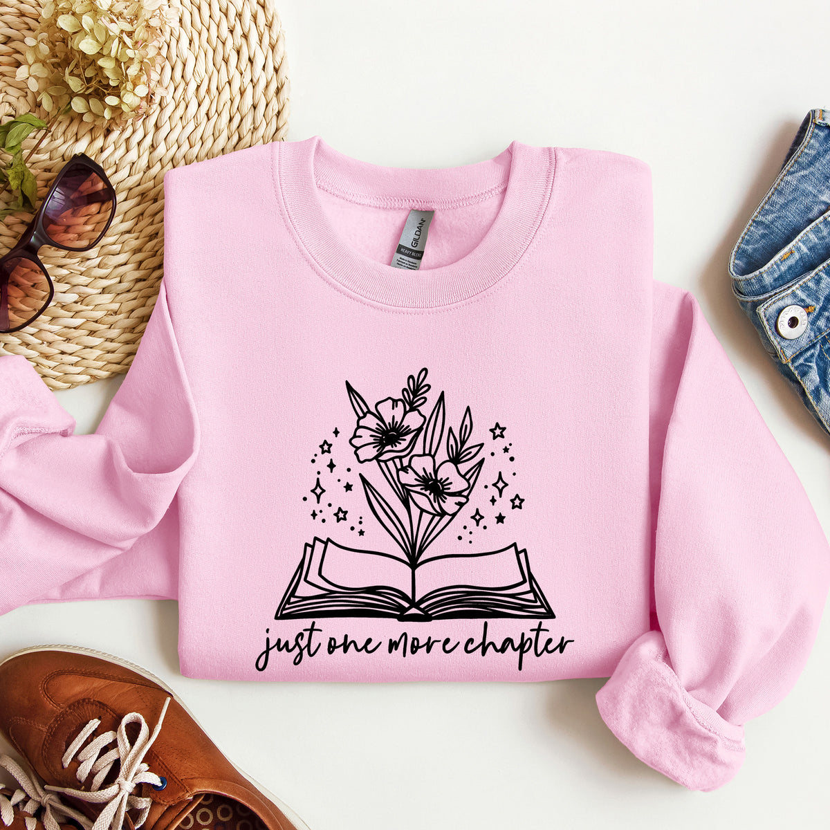A folded light pink sweatshirt with a black graphic of an open book and flowers, styled in a flat lay with a straw hat, sunglasses, jeans, and a brown shoe on a white background.