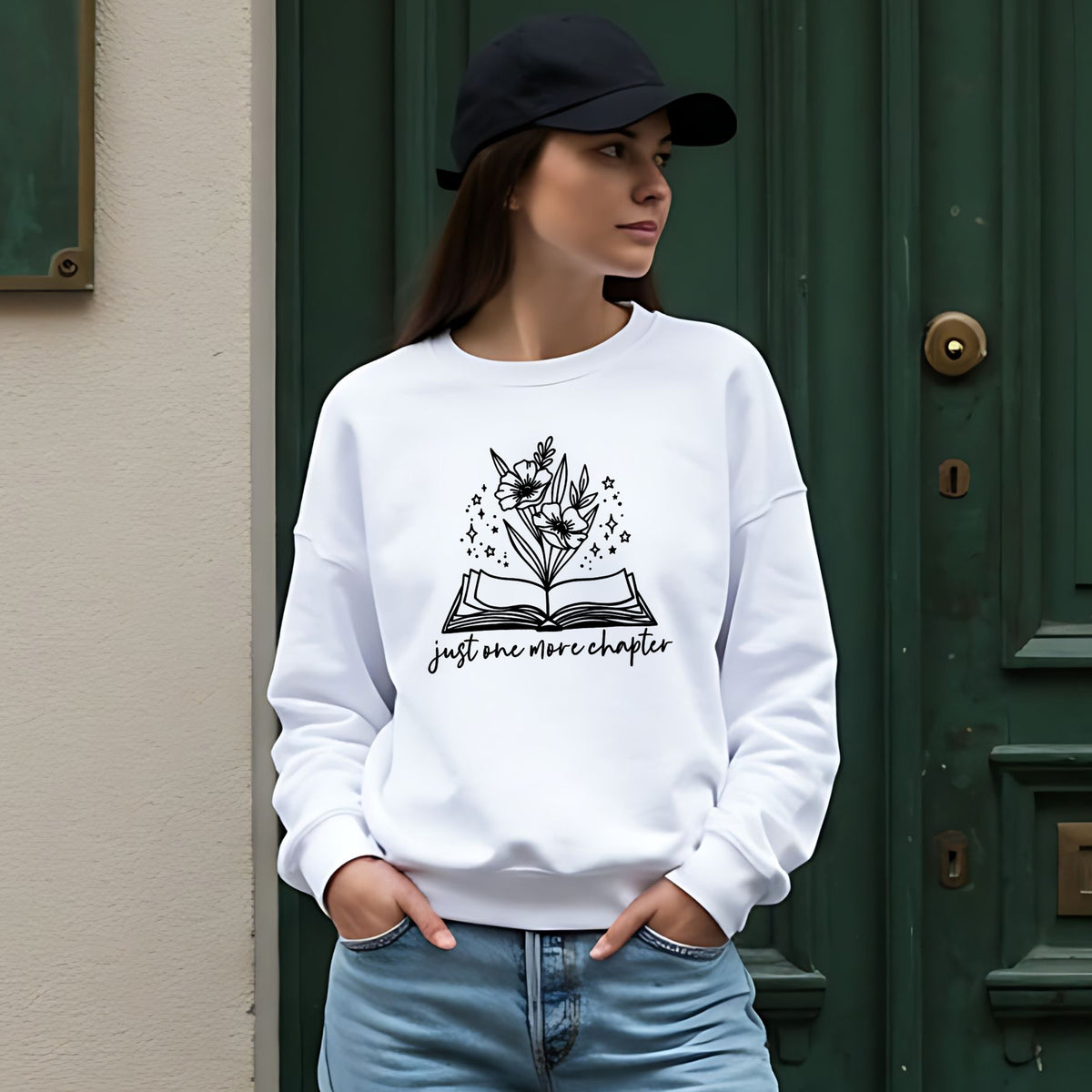 A woman wearing a white crewneck sweatshirt with a black graphic of an open book and flowers, with the text 'just one more chapter', paired with a black baseball cap and light blue jeans, standing in front of a dark green door.