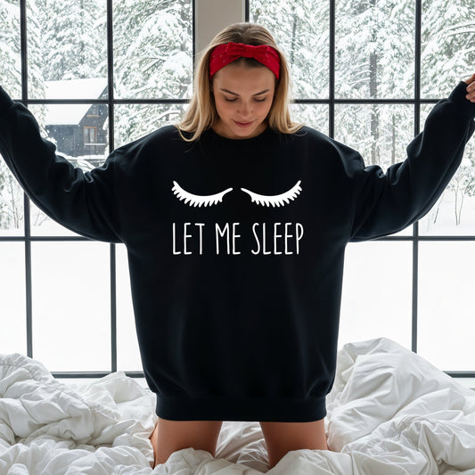 A woman wearing a black crewneck sweatshirt with a white graphic of eyelashes and the text 'LET ME SLEEP' while kneeling on a white bed in front of a window with a snowy outdoor view.