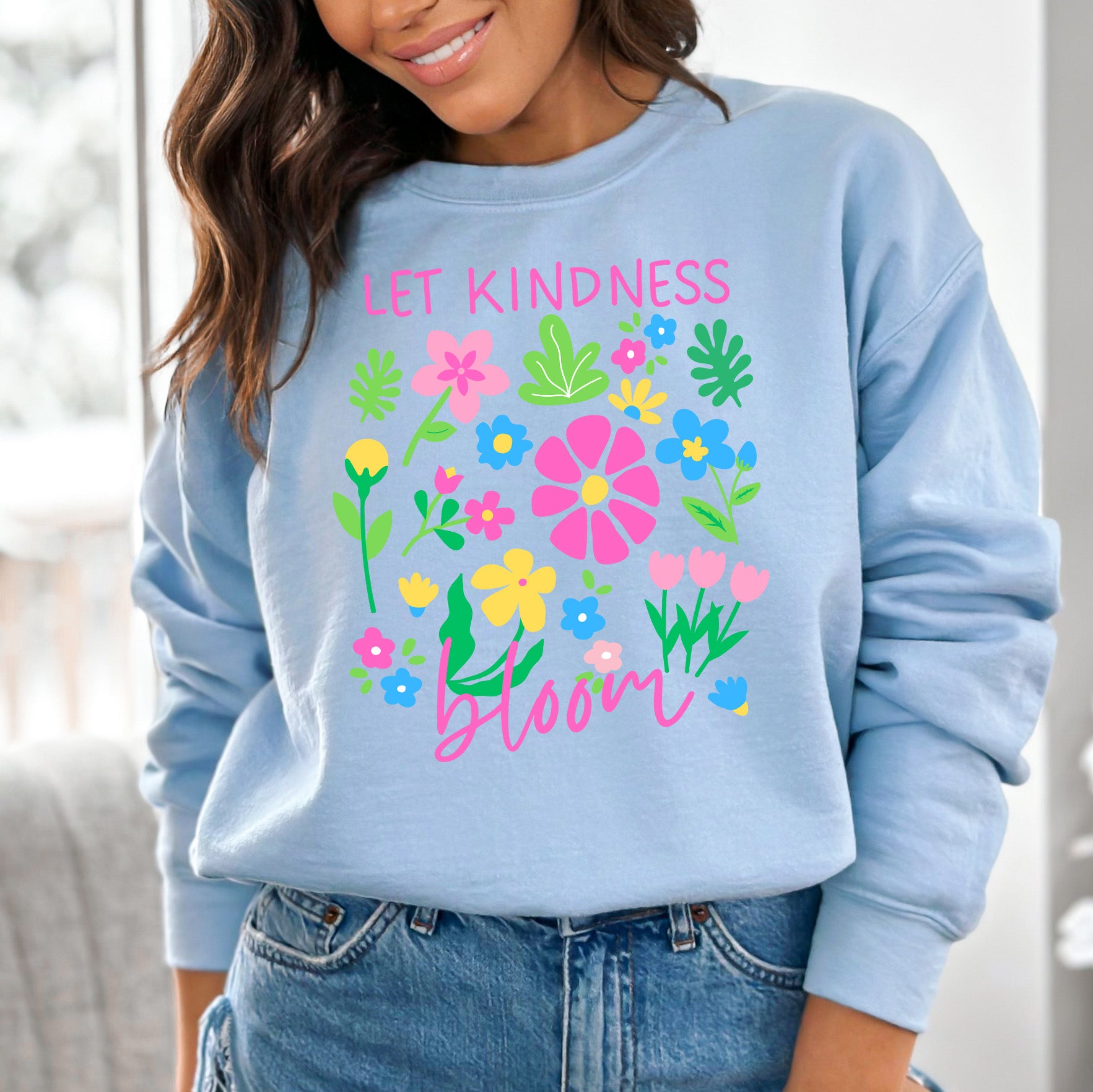 Light blue sweatshirt with colorful floral design and text 'Let kindness bloom' worn by a person.