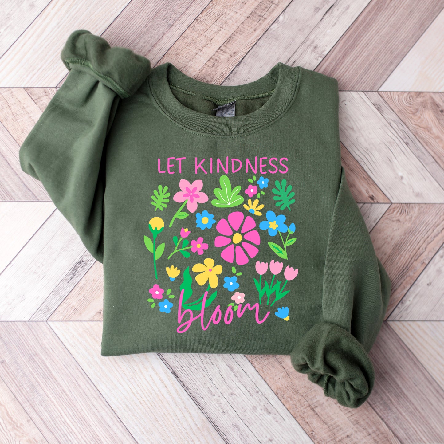 Green sweatshirt with floral design and 'Let Kindness Bloom' text on a wooden floor.