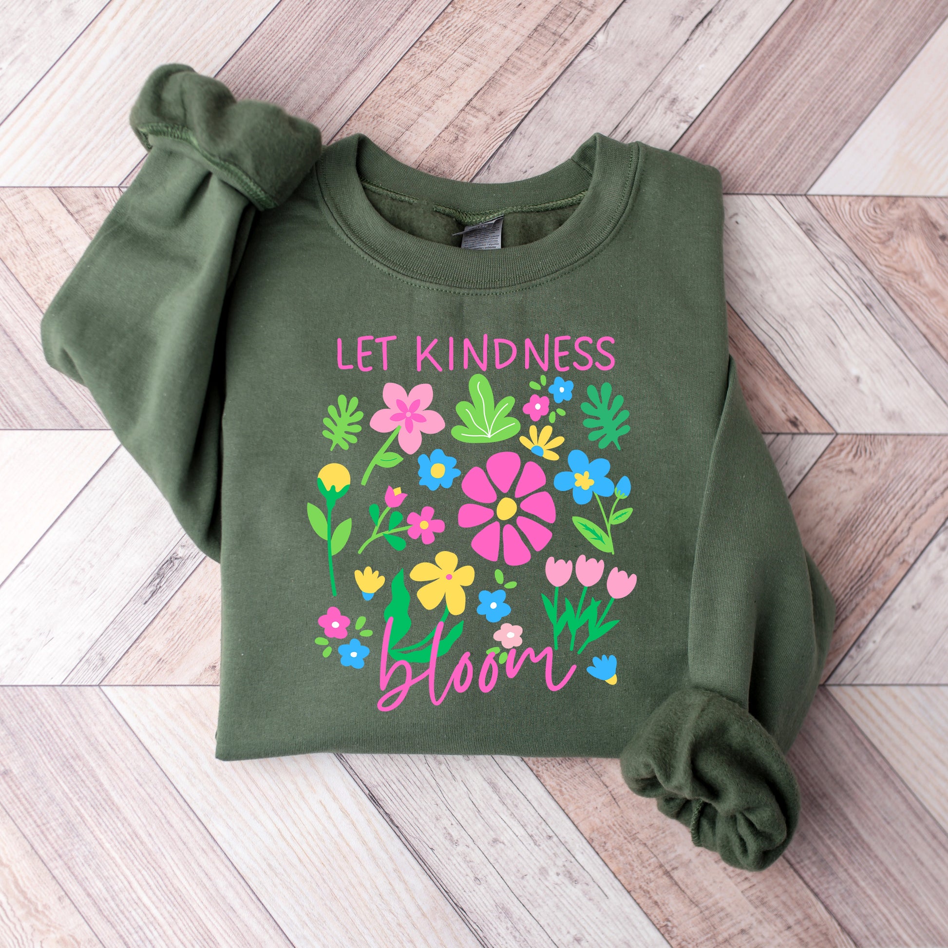 Green sweatshirt with floral design and 'Let Kindness Bloom' text on a wooden floor.