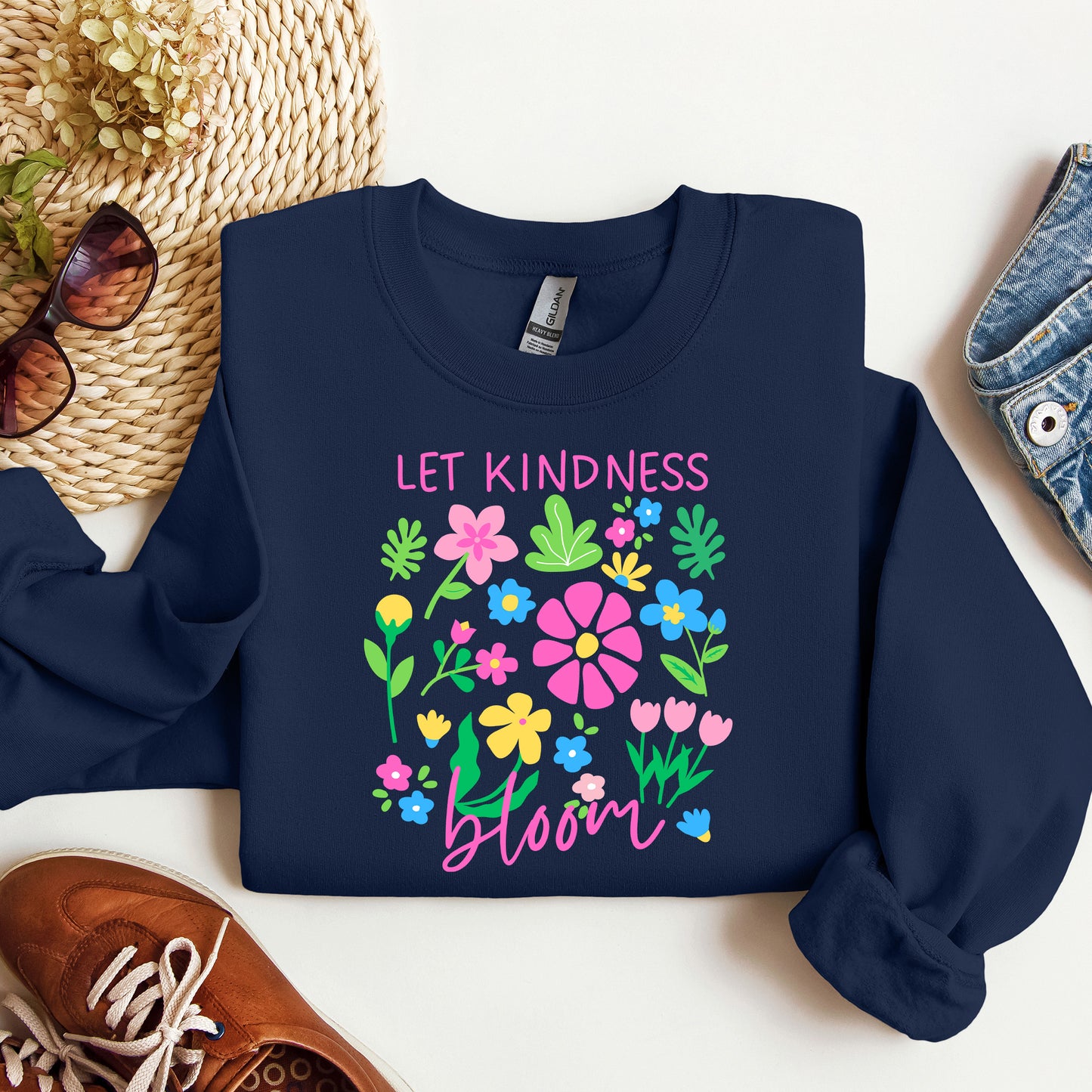 Navy blue sweatshirt with floral design and 'Let Kindness Bloom' text on a white surface.