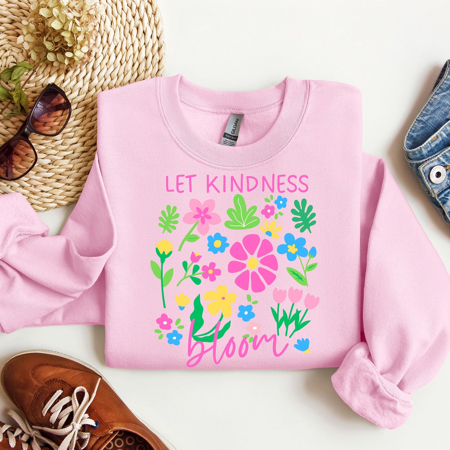 Pink sweatshirt with floral design and 'Let Kindness Bloom' text on a white surface with accessories.