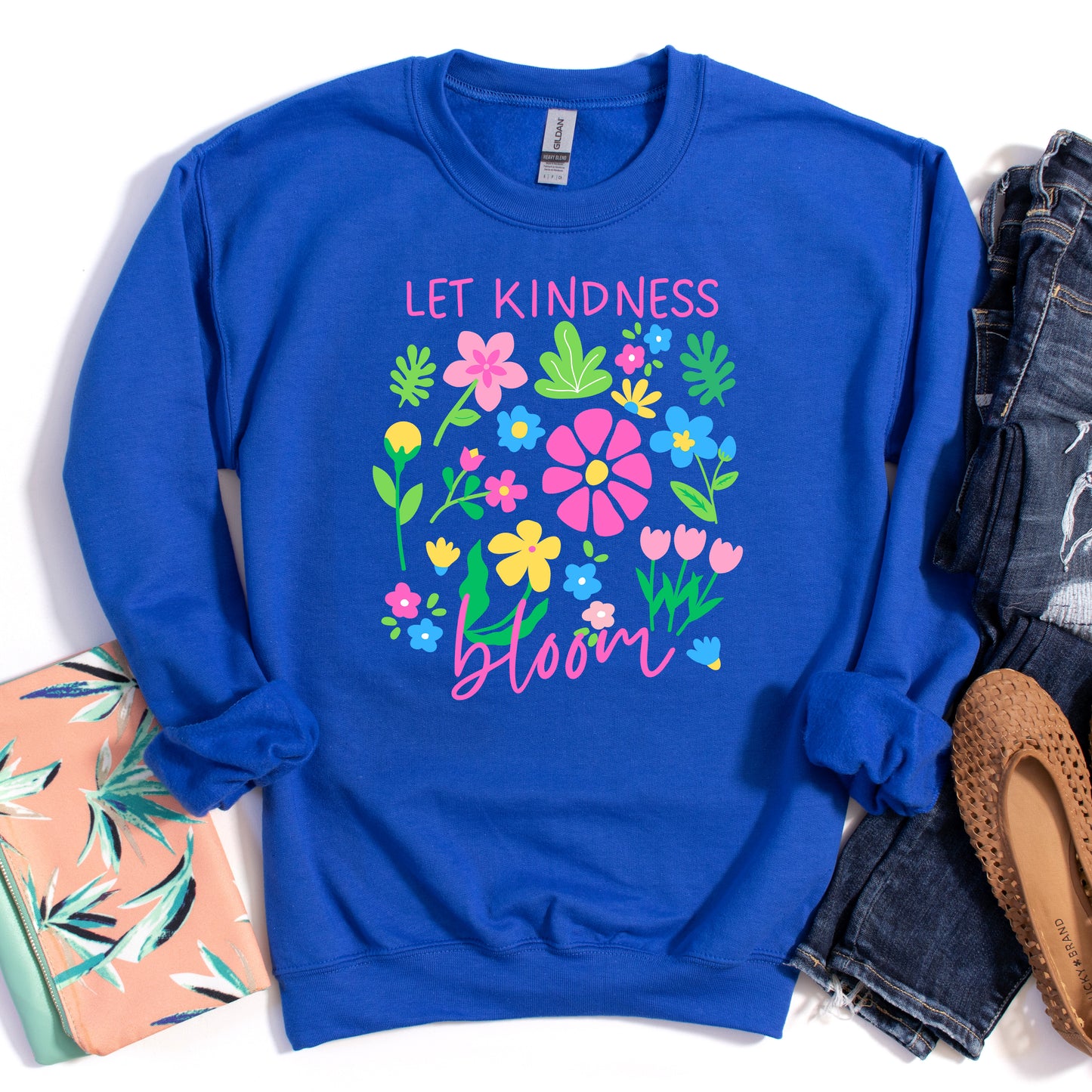Blue sweatshirt with floral design and 'Let Kindness Bloom' text, surrounded by casual items on a white background.