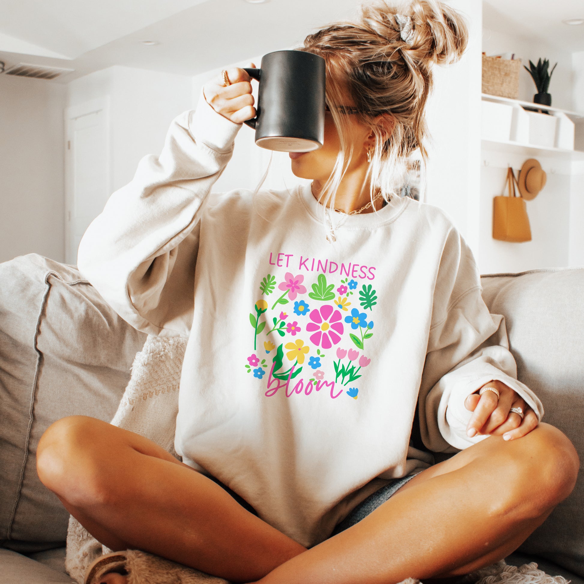 Woman wearing a white sweatshirt with colorful text and flowers, sitting on a couch holding a mug.