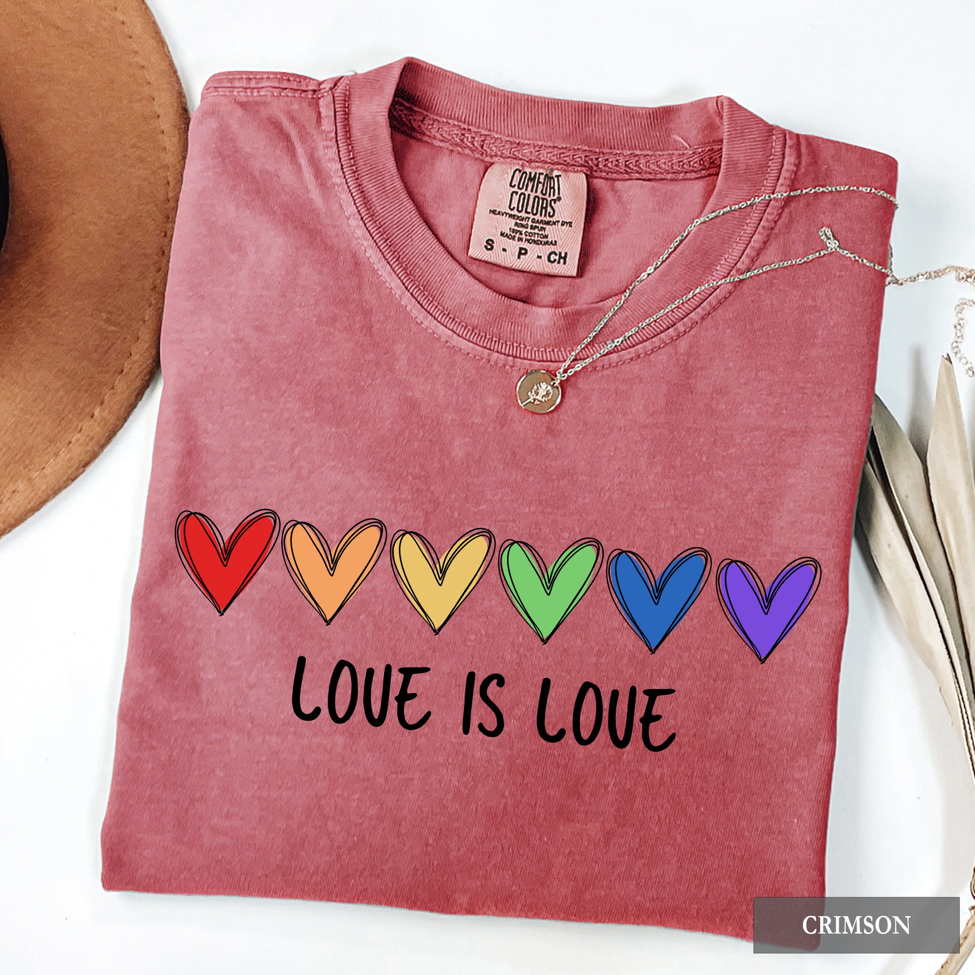 Folded pink t-shirt with rainbow hearts and 'LOVE IS LOVE' text, next to a hat and necklace on a white background.