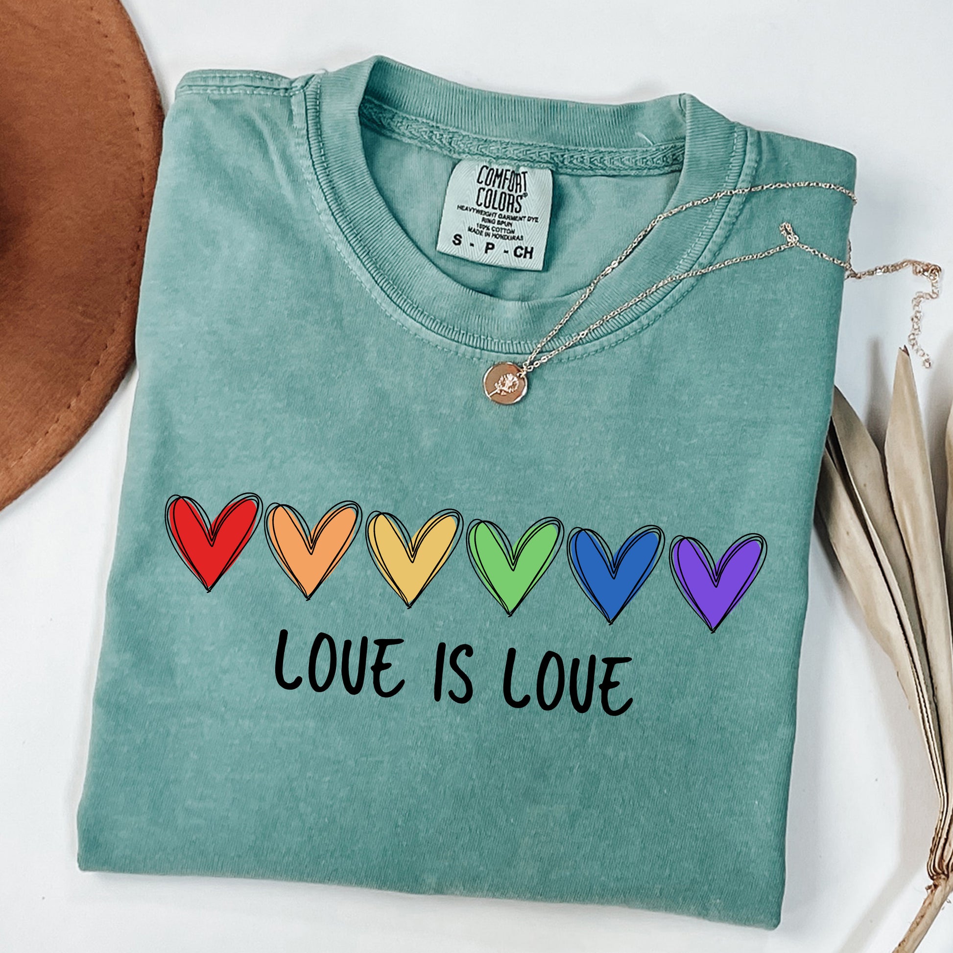 Folded teal t-shirt with rainbow hearts and 'Love is Love' text on a white background