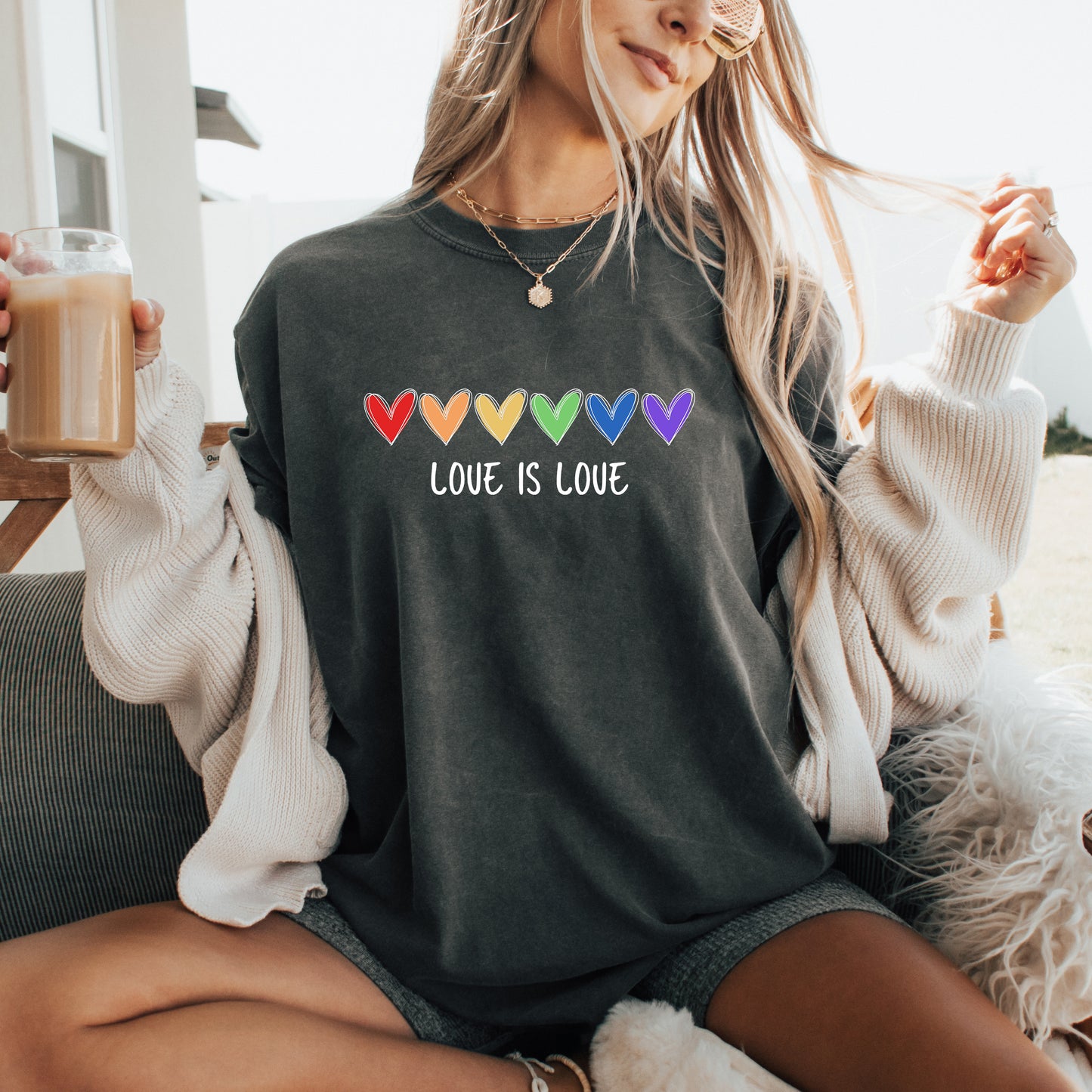 Person wearing a dark gray sweatshirt with 'Love is Love' text and rainbow hearts, holding a drink.