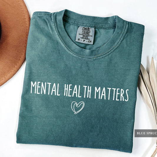 A folded slate blue-green t-shirt with 'MENTAL HEALTH MATTERS' and a heart outline printed in white on the front. The shirt is styled on a white surface with a brown hat and dried leaves, with a 'BLUE SPRUCE' brand tag visible on the bottom right.
