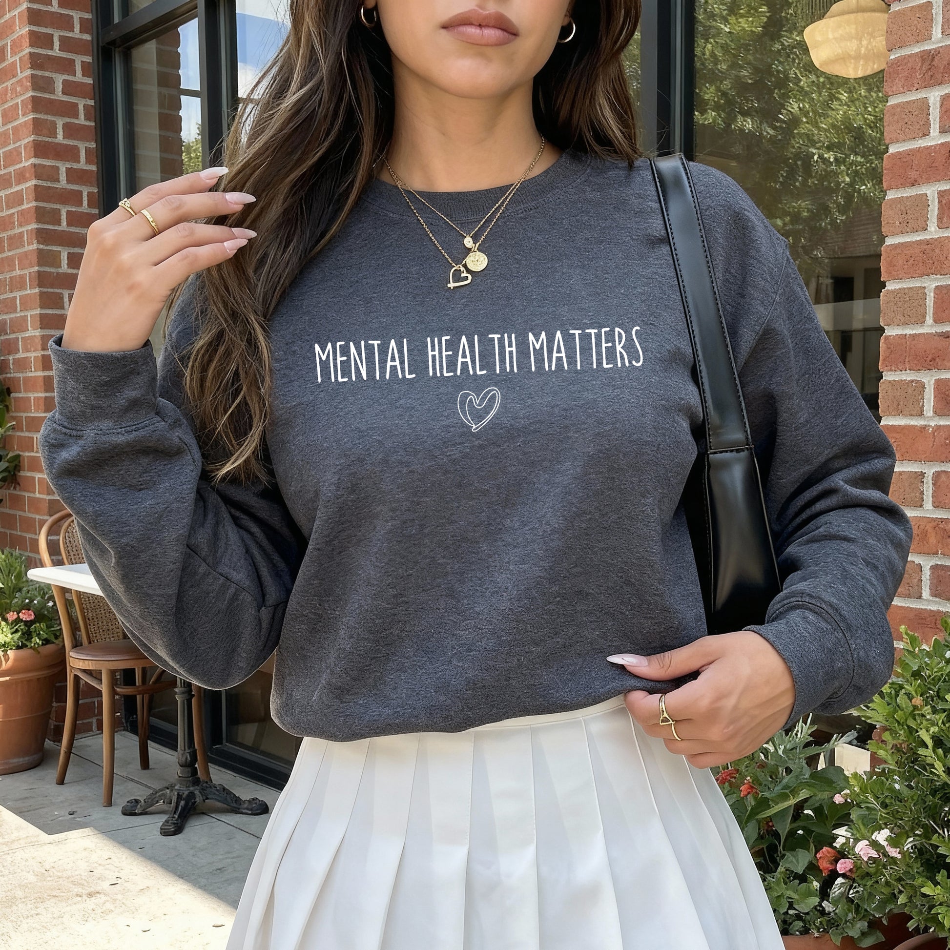 A woman wearing a dark gray crewneck sweatshirt with the text 'MENTAL HEALTH MATTERS' and a small heart outline printed on the front, paired with a white pleated skirt. She is standing outdoors in front of a brick building.