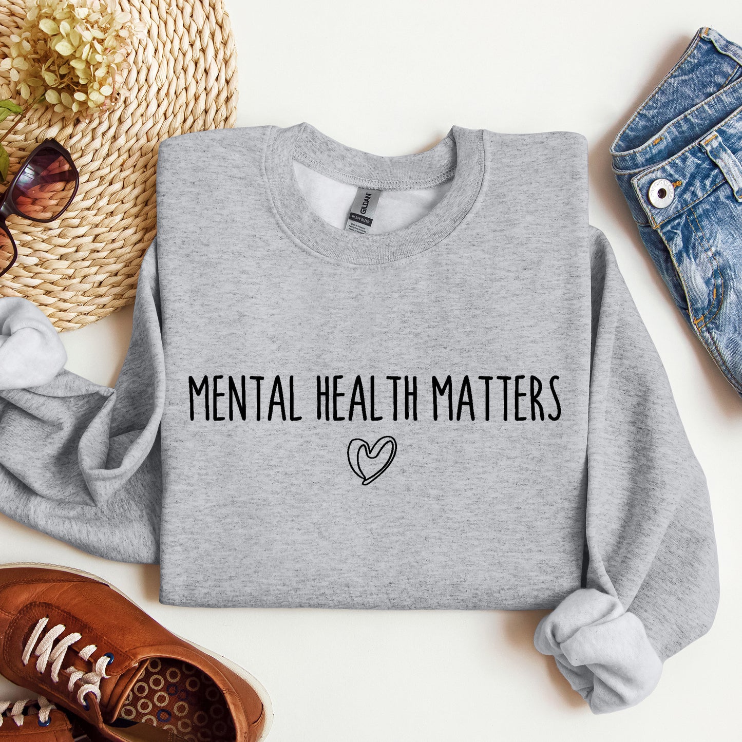 A folded heather gray sweatshirt with the words 'MENTAL HEALTH MATTERS' and a small heart outline printed on the front, styled in a flat lay with jeans, sunglasses, a woven hat, shoes, and dried flowers on a white background.