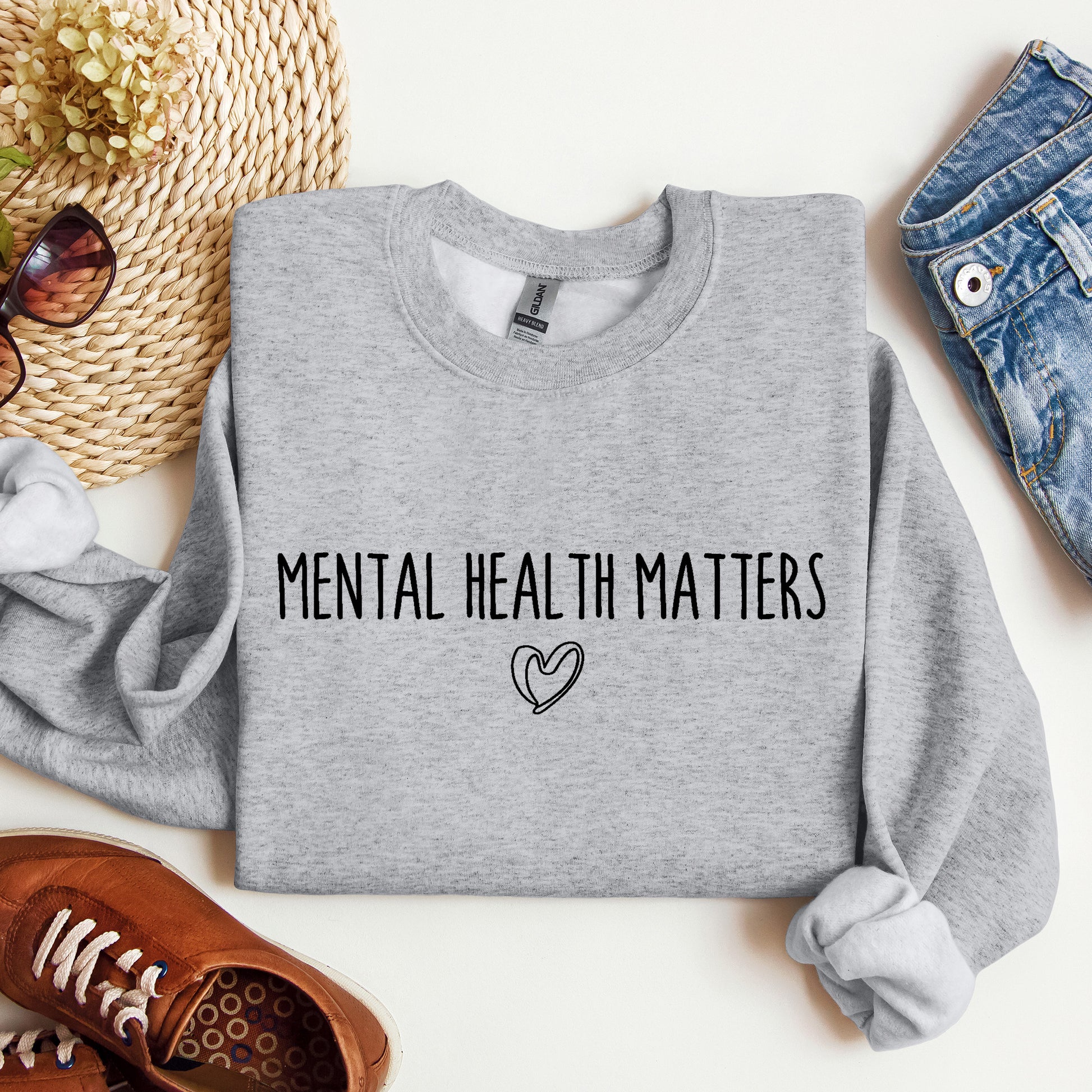 A folded heather gray sweatshirt with the words 'MENTAL HEALTH MATTERS' and a small heart outline printed on the front, styled in a flat lay with jeans, sunglasses, a woven hat, shoes, and dried flowers on a white background.