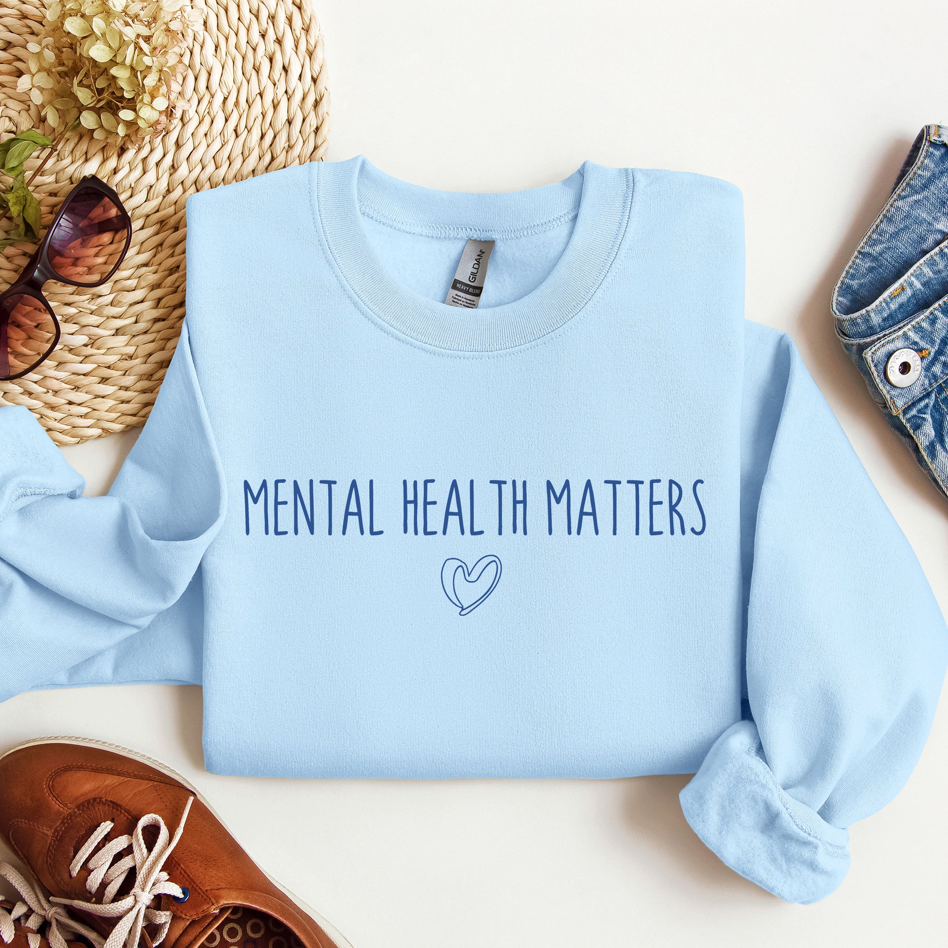 A light blue crewneck sweatshirt with the text 'MENTAL HEALTH MATTERS' and a small heart outline printed on the front. The sweatshirt is styled in a flat lay with a straw hat, sunglasses, a brown shoe, and a denim pocket on a white background.