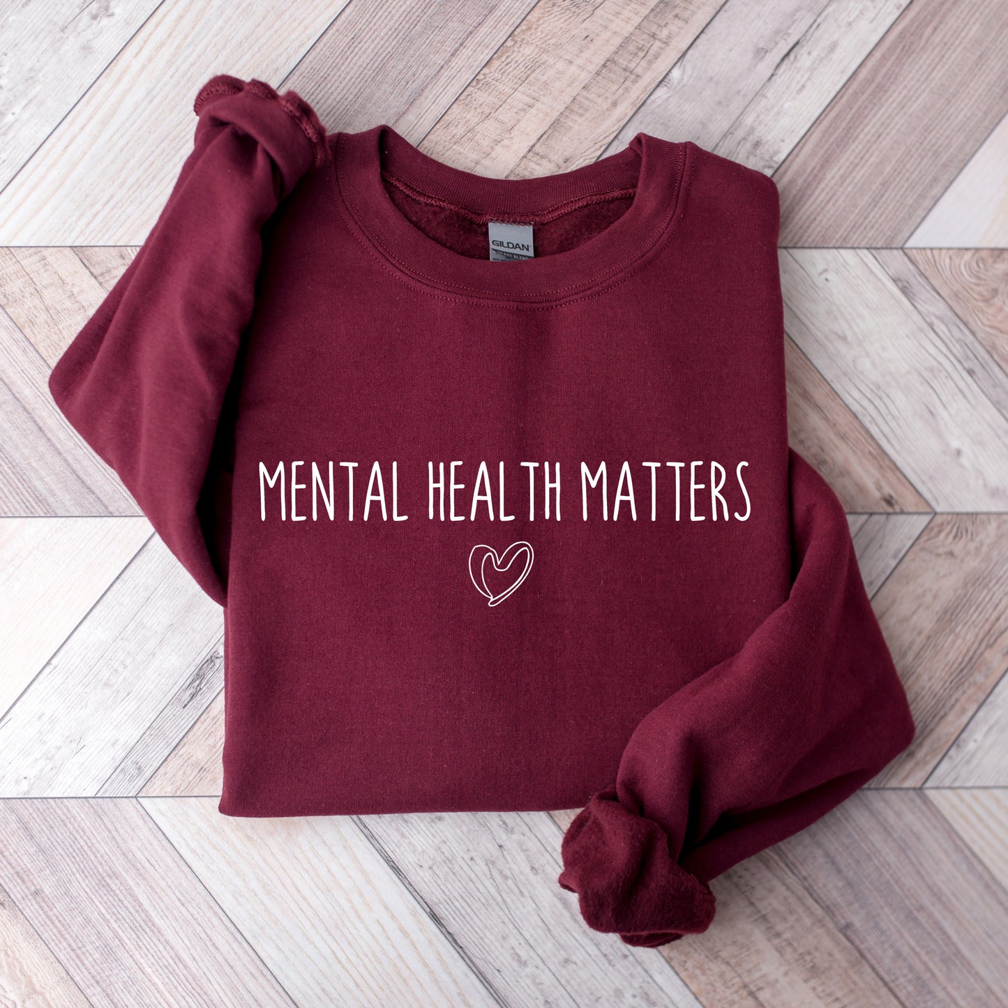 A folded maroon sweatshirt with the words 'MENTAL HEALTH MATTERS' and a small heart outline printed in white on the front, laid flat on a light wood plank background.