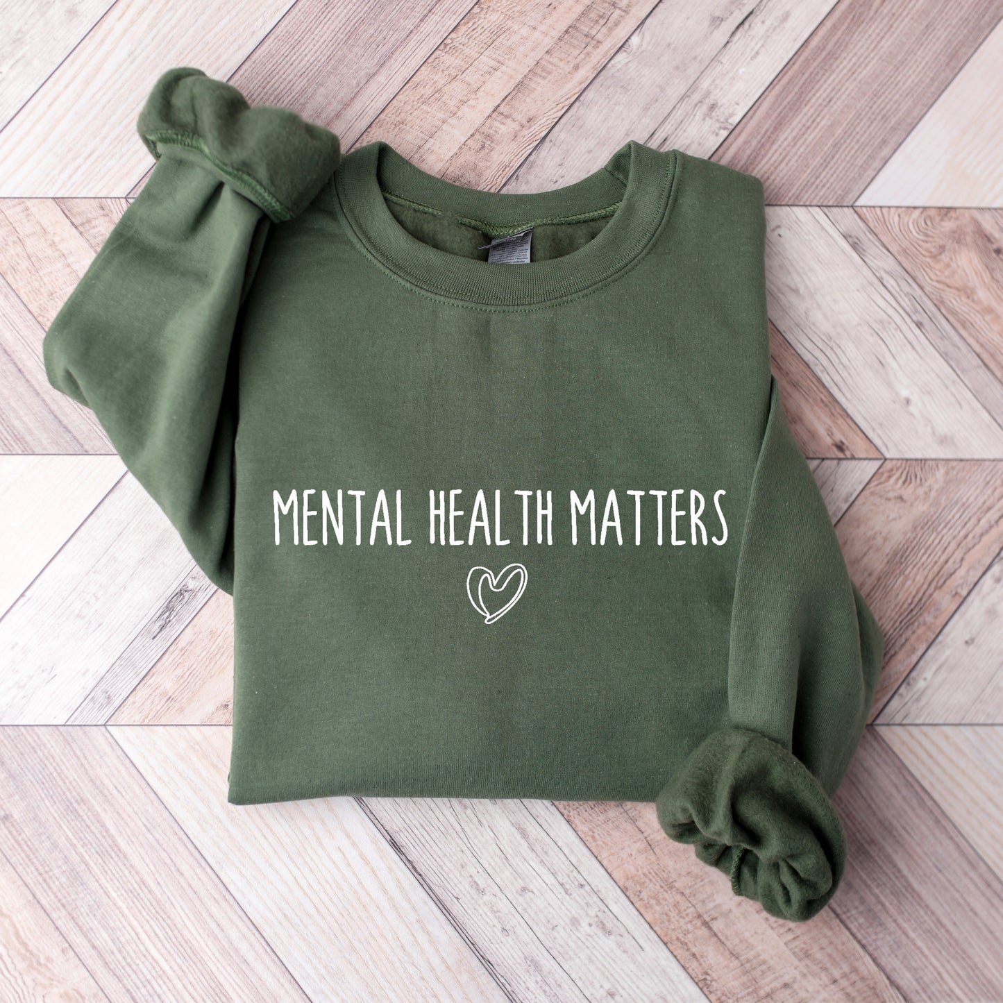 An olive green long-sleeve sweatshirt with 'Mental Health Matters' printed on the front, laid flat on a light wood plank background.