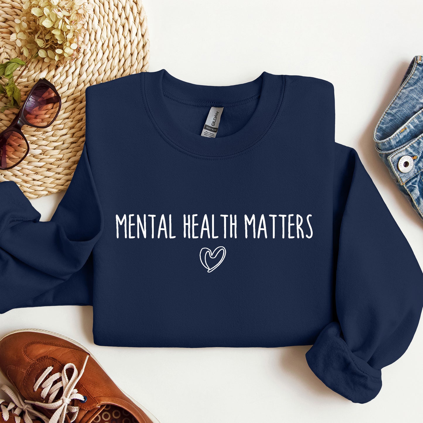 A navy blue long-sleeve crewneck sweatshirt with the phrase 'Mental Health Matters' printed in white across the chest. The sweatshirt is styled in a flat lay with a woven straw hat, sunglasses, a brown leather shoe, and a denim jacket on a white background.
