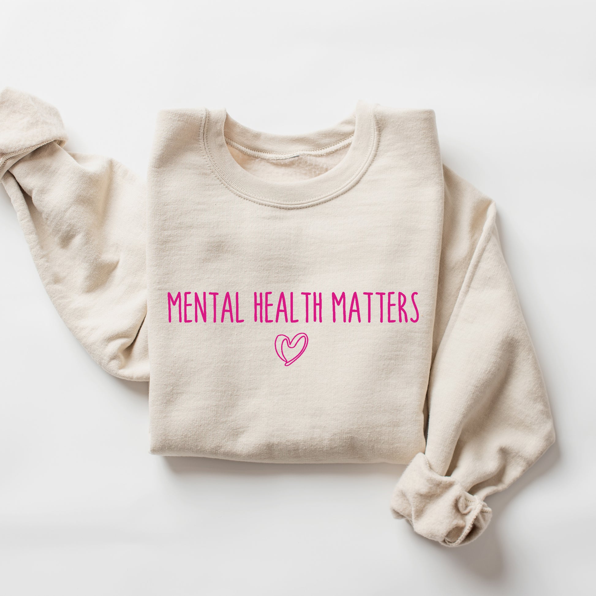 A folded cream-colored sweatshirt with the words 'MENTAL HEALTH MATTERS' printed in bright pink across the chest, accompanied by a small pink heart outline below, displayed on a clean white background.