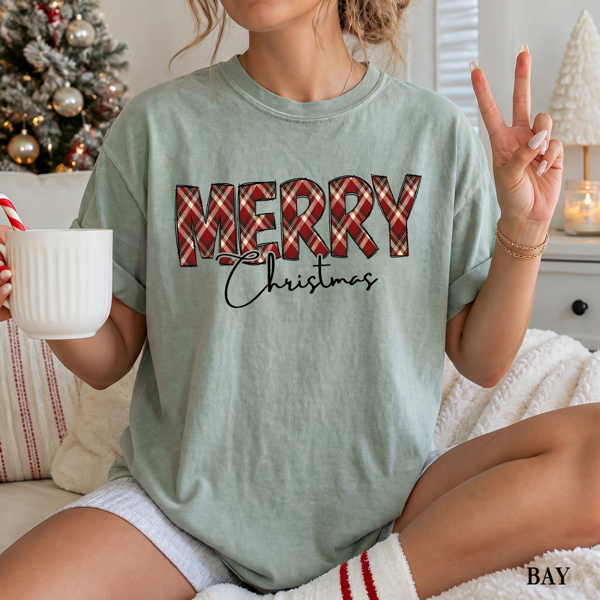 A woman wearing a bay Comfort Colors shirt with Merry in red, green and cream plaid and Christmas underneath it.