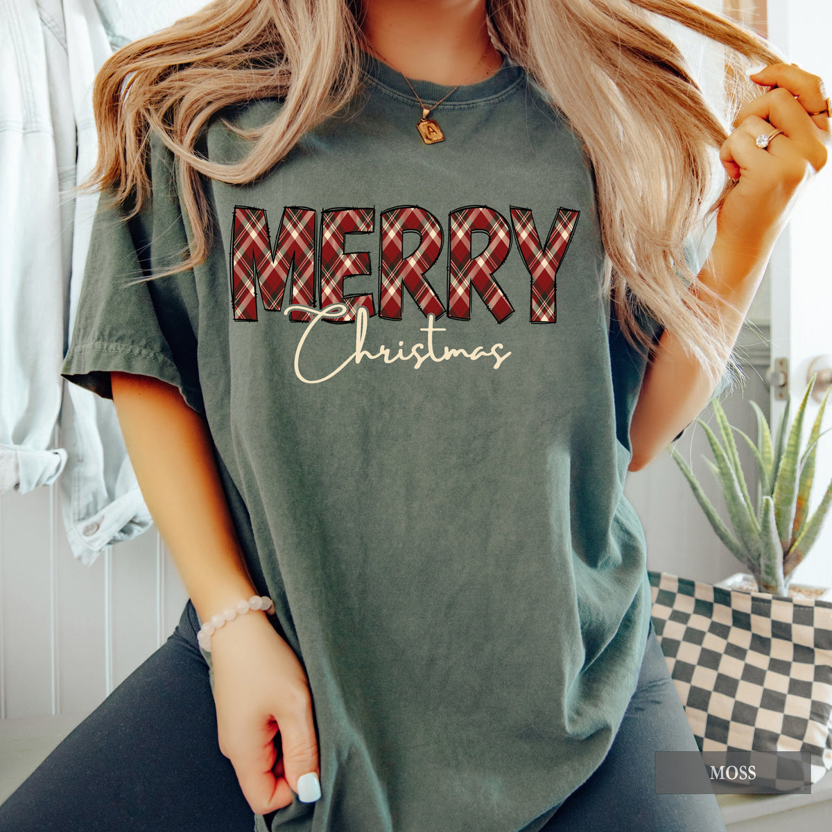 A woman wearing a moss Comfort Colors shirt with Merry in red, green and cream plaid and Christmas underneath it.