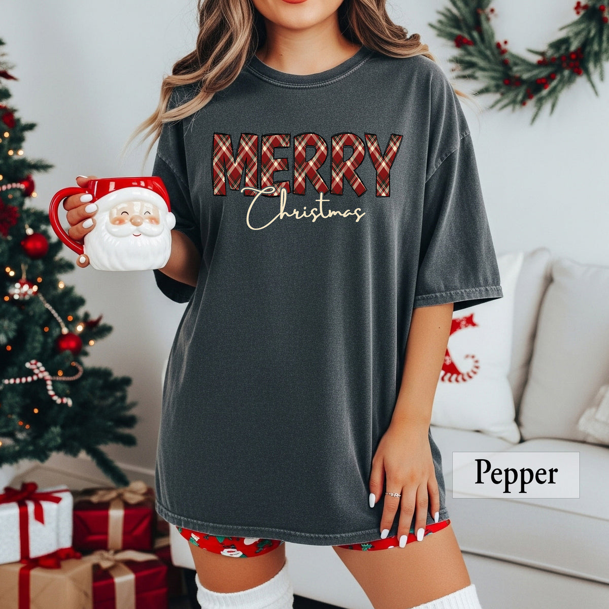 A woman wearing a pepper Comfort Colors shirt with Merry in red, green and cream plaid and Christmas underneath it.